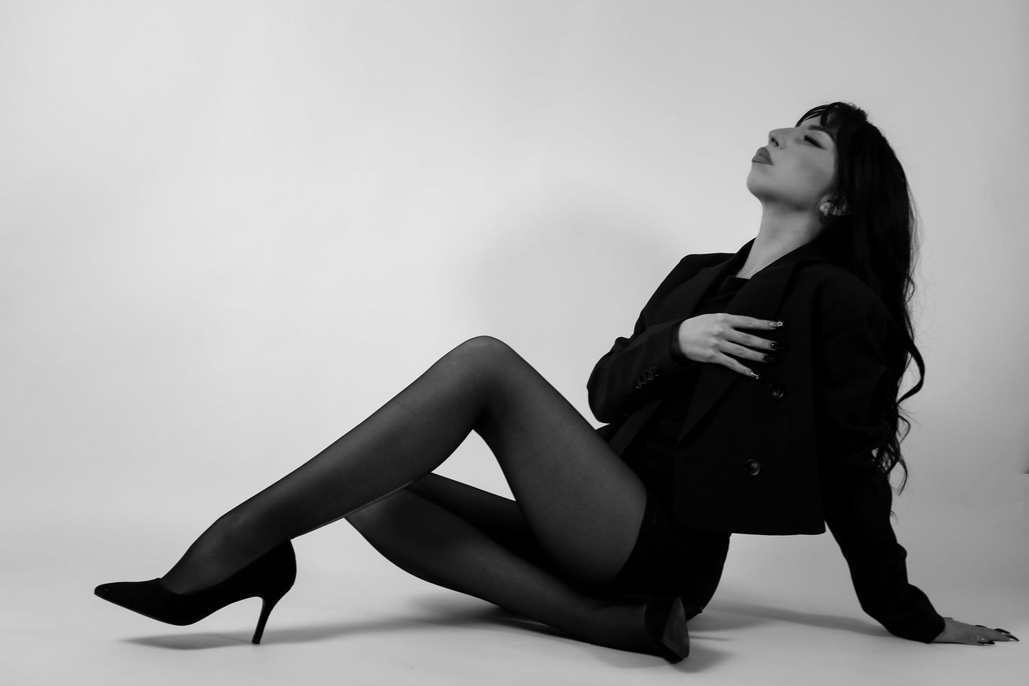 Black and white photo of a woman in a black blazer and heels sitting gracefully on a studio floor.