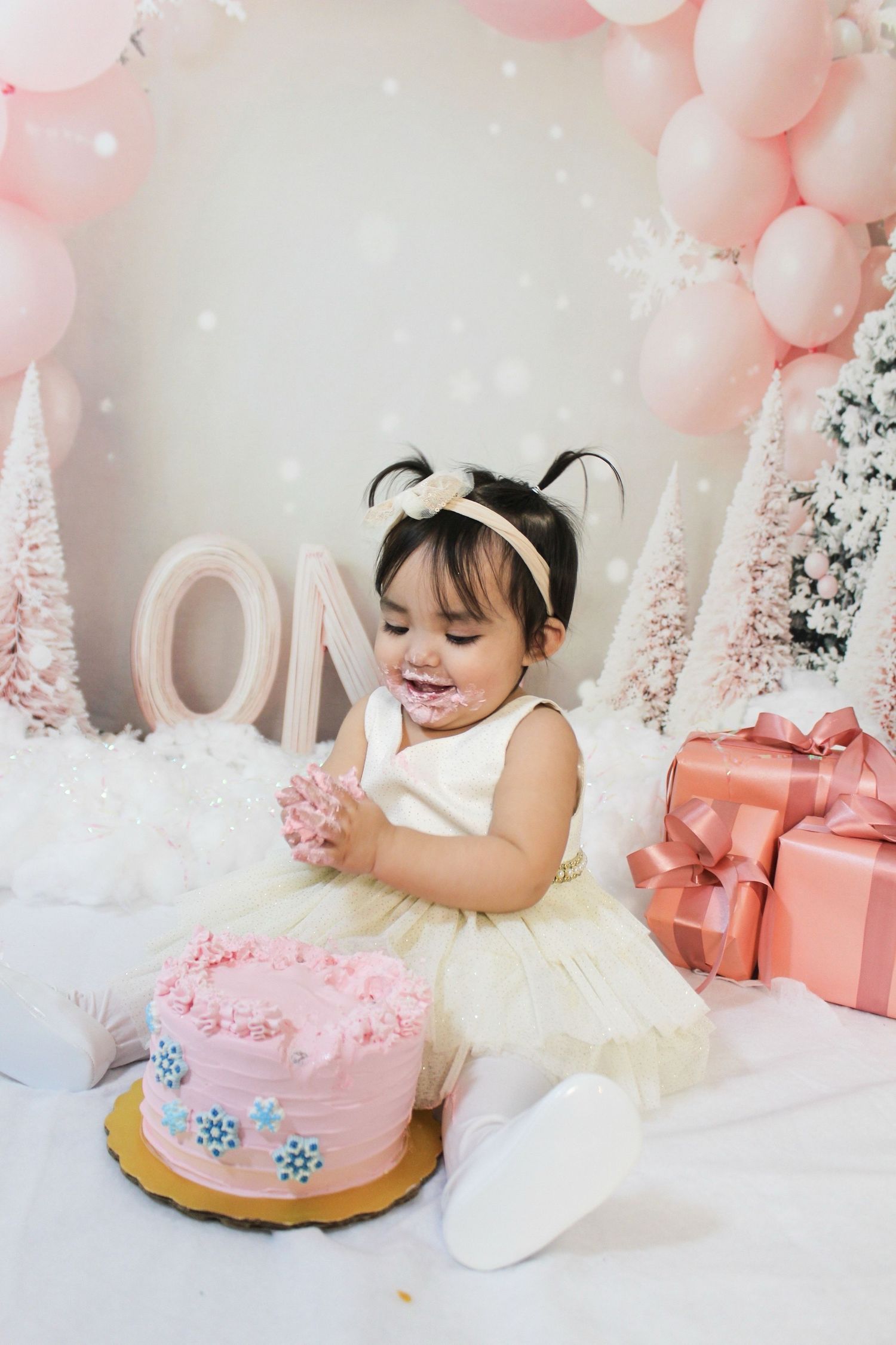 Adorable baby girl in white tutu dress smiling at pink birthday cake during first birthday cake smash photo session.