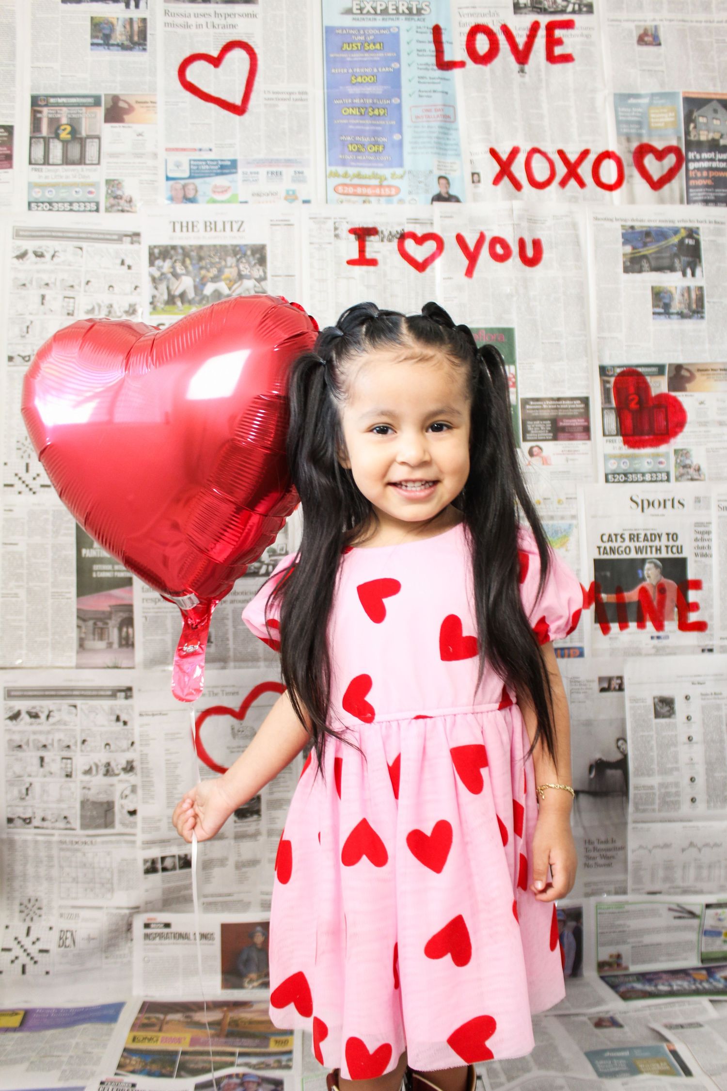 A smiling little girl in a pink heart dress holds a red heart balloon against a Valentine's Day newspaper backdrop.