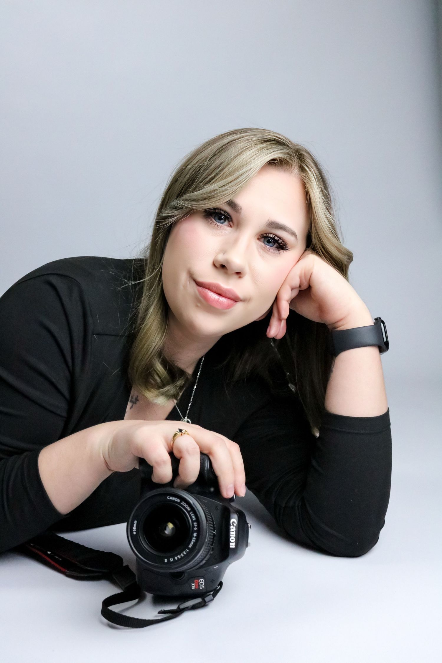 Smiling blonde female photographer leaning on white surface holding Canon DSLR camera with hand on cheek.