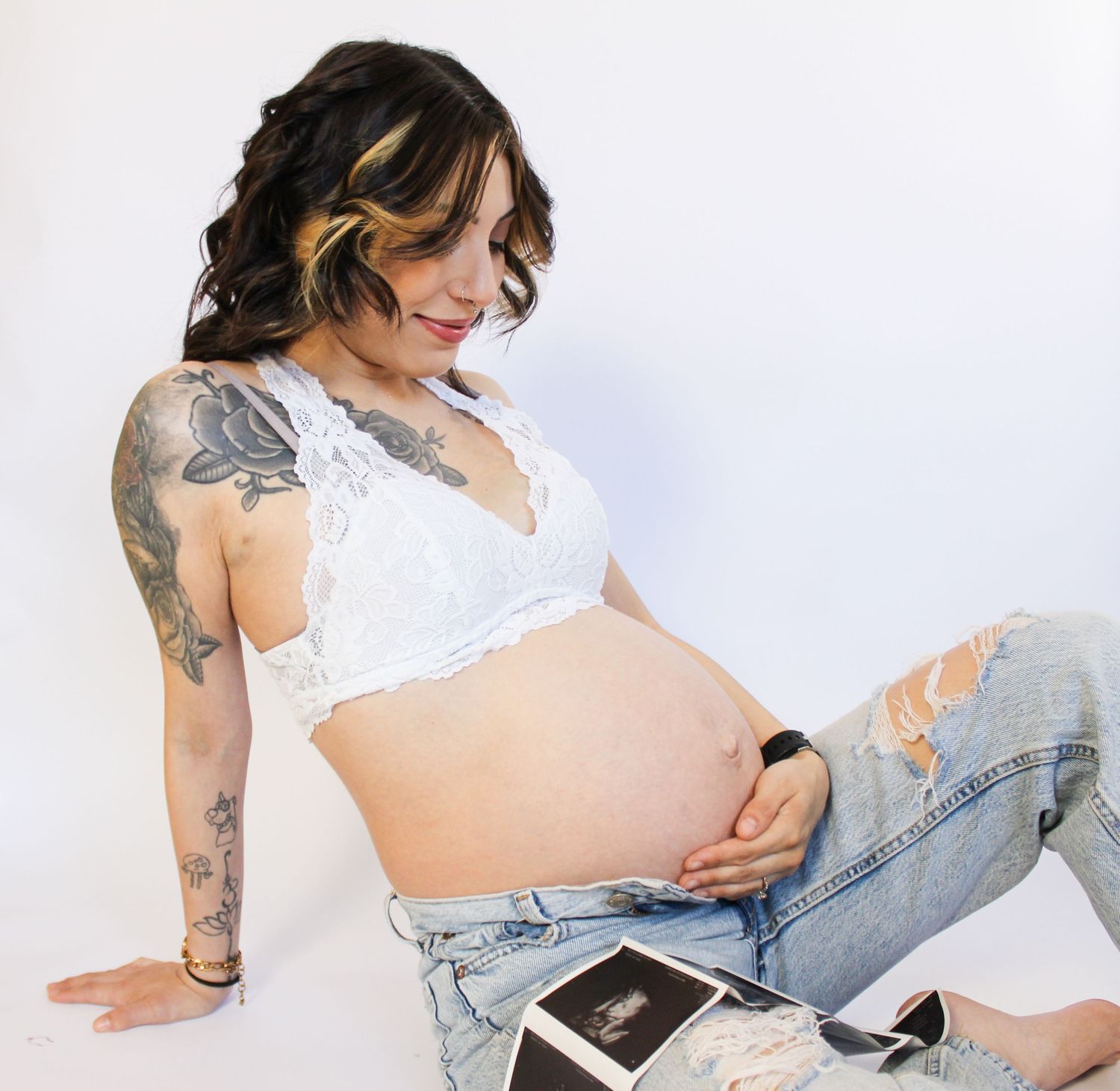 Tattooed pregnant woman in white lace bra and jeans sitting with ultrasound photos while holding her baby bump.