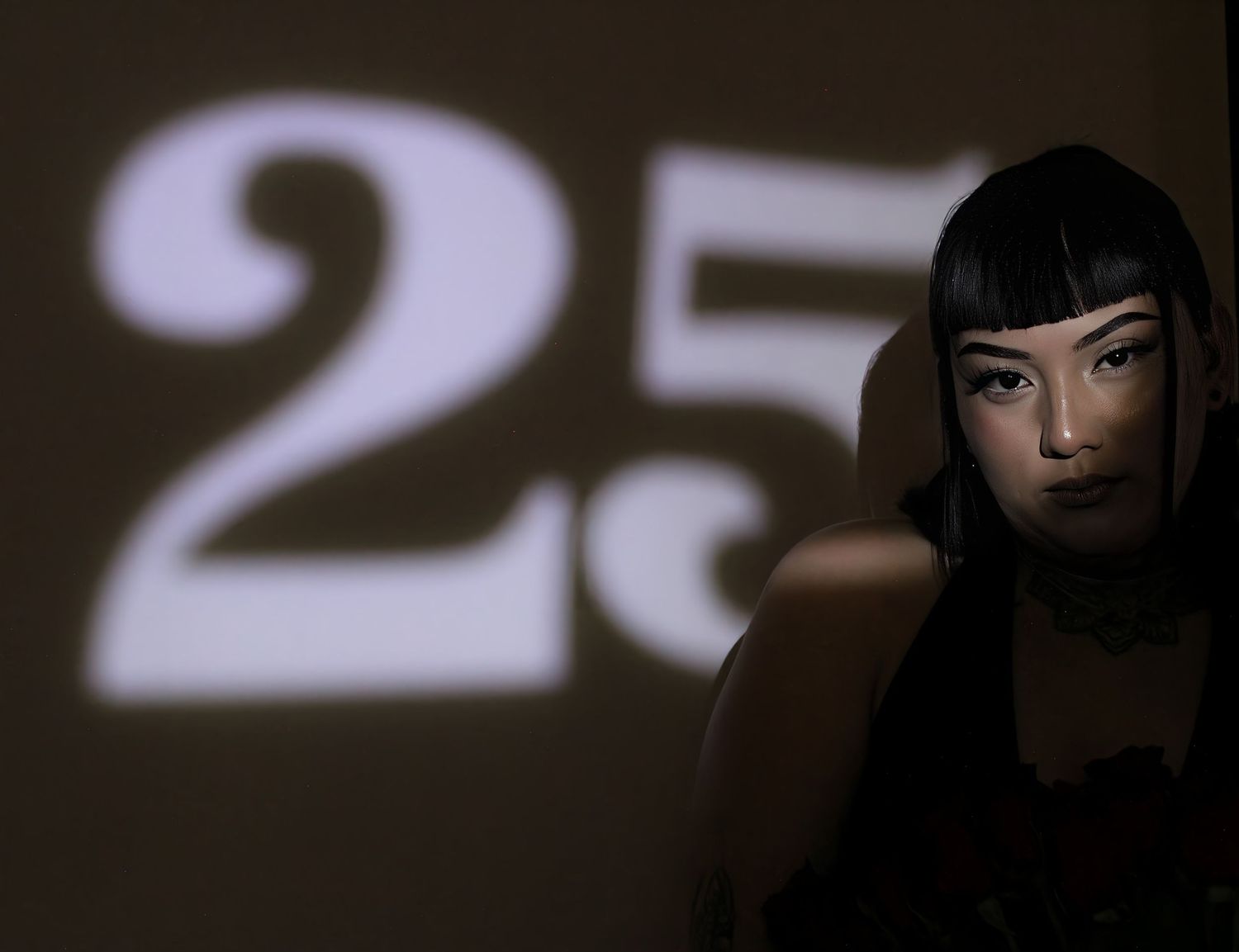 Woman with blunt bangs posing in dramatic shadow with a large number 25 projected on the wall behind her.