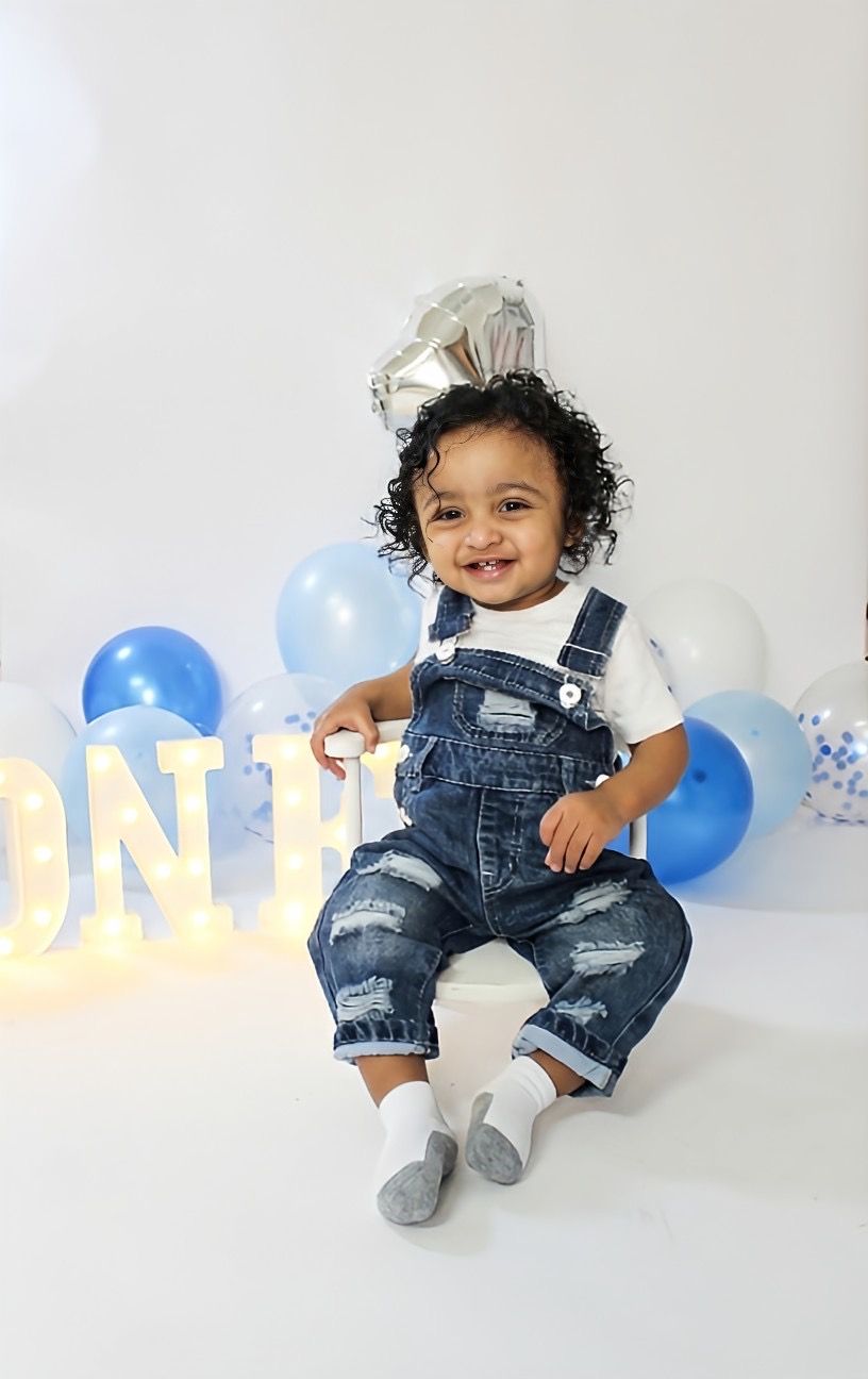 Smiling toddler in denim overalls sitting near ONE sign and blue balloons for first birthday photo.