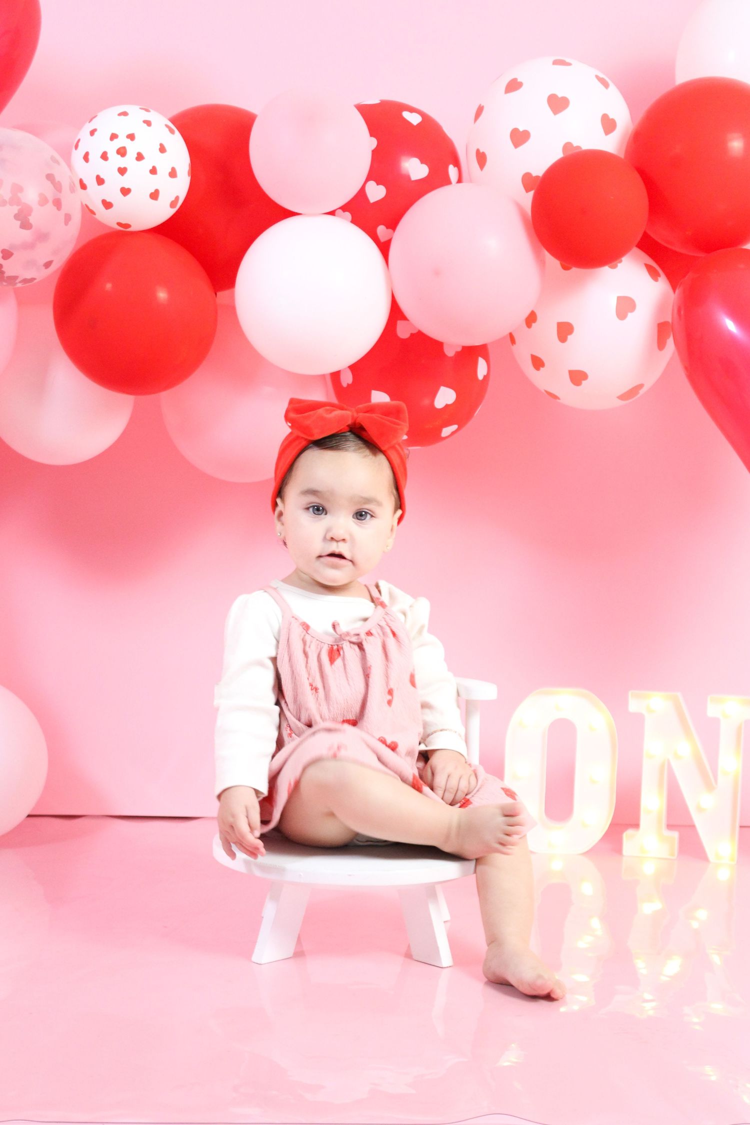 Baby girl in pink outfit sitting on white chair at Valentine's Day themed first birthday session.