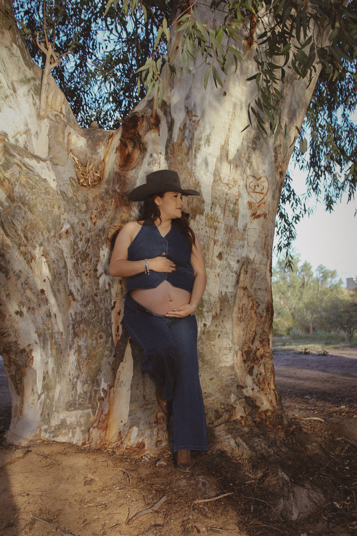 Pregnant woman in navy blue outfit and cowboy hat leaning against large eucalyptus tree outdoors.