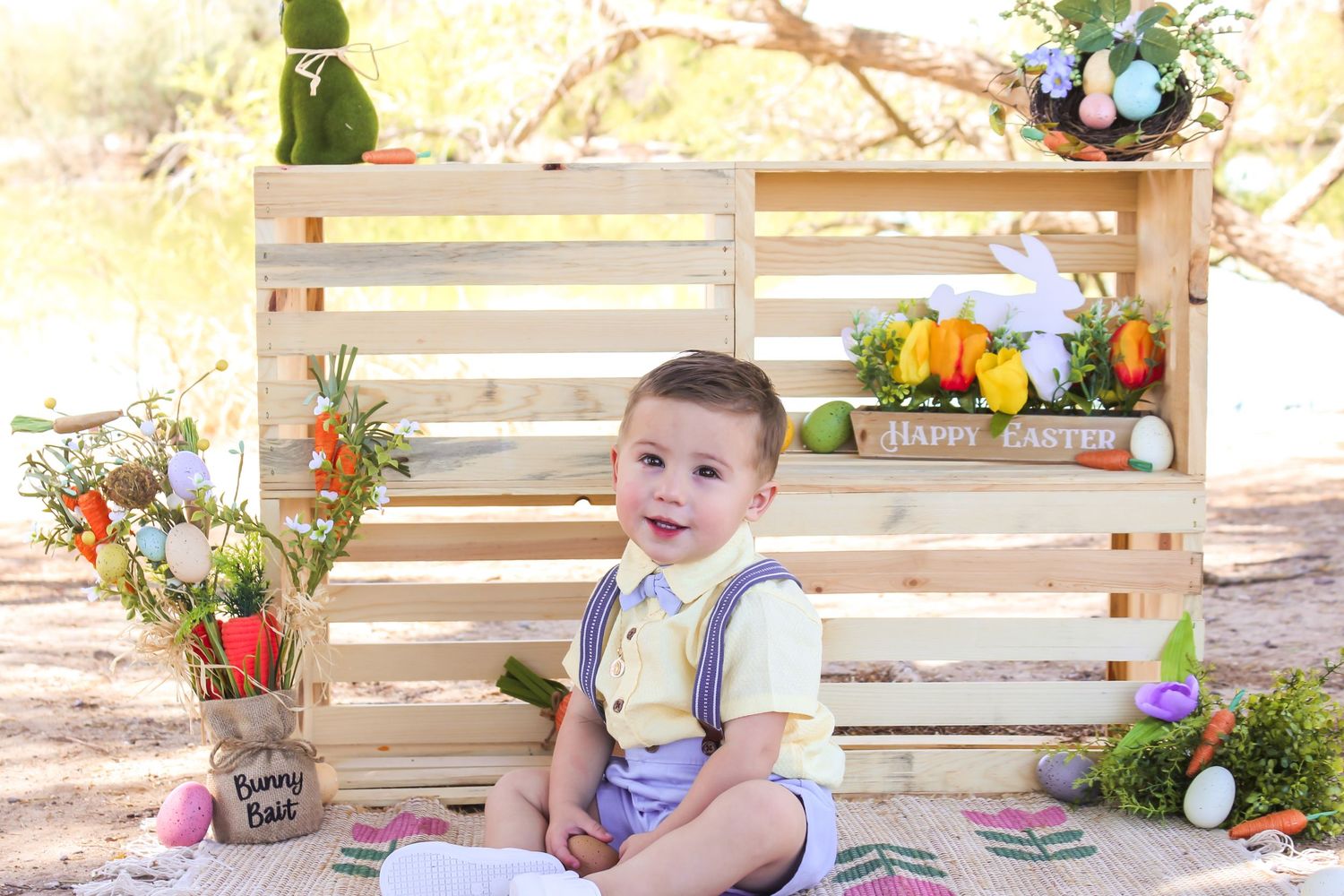Adorable toddler boy in suspenders sitting at a colorful Easter-themed outdoor photo setup with spring flowers.