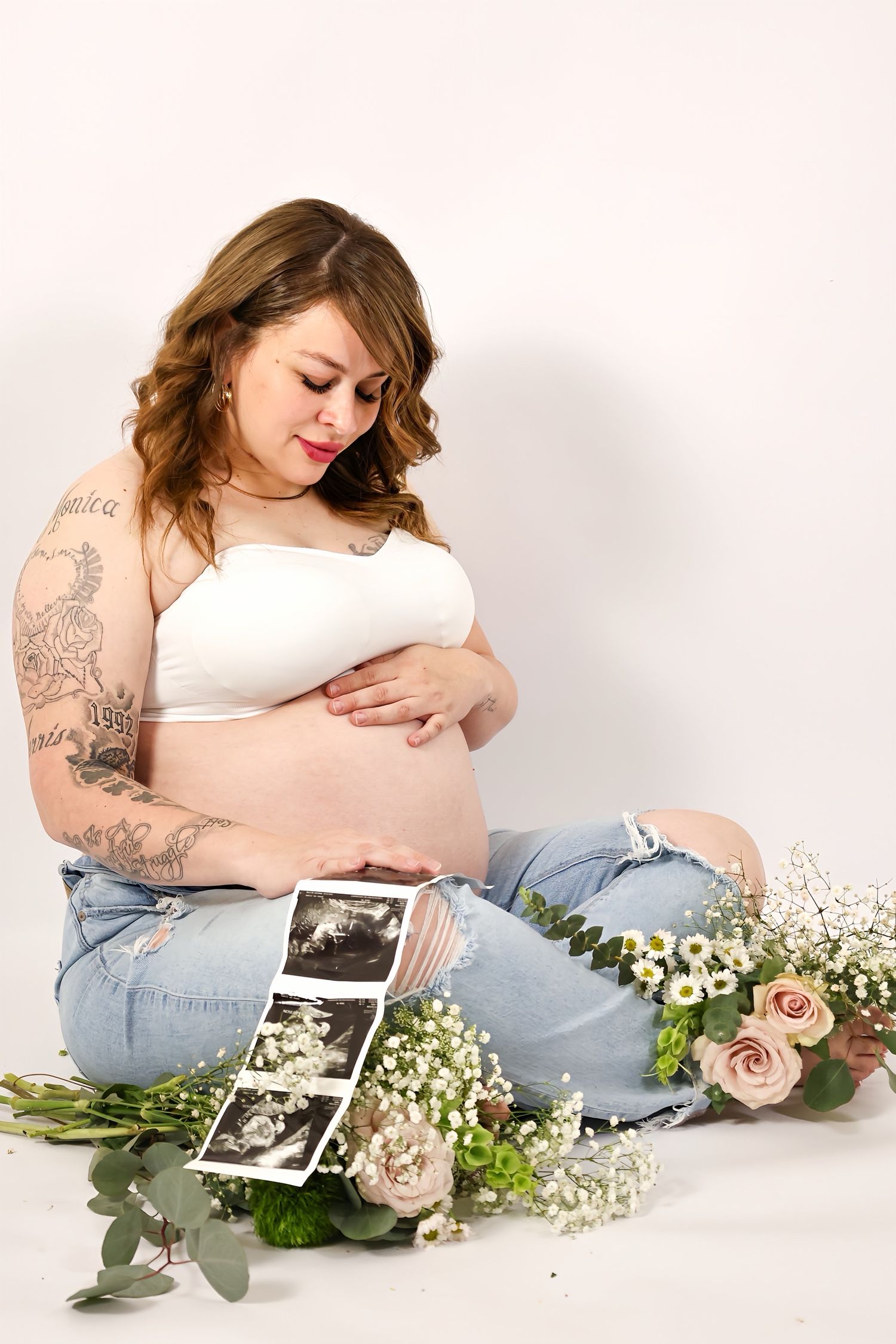 Tattooed pregnant woman in white crop top holds ultrasound photos surrounded by fresh flowers.