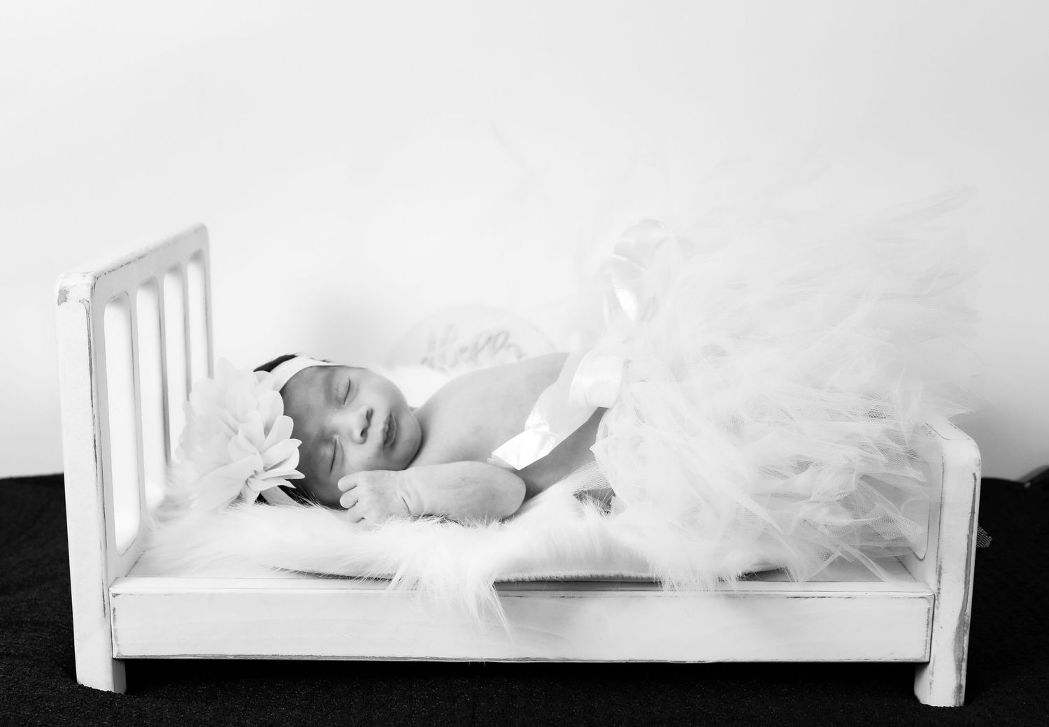Adorable newborn baby in white tutu lying on a small white wooden bed in black and white photo.