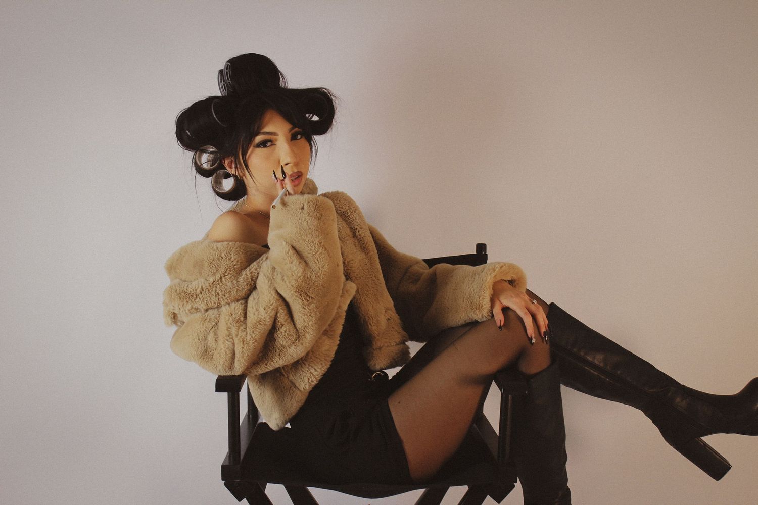 Woman in fur coat and knee-high boots posing on chair with hair in rollers against white background.