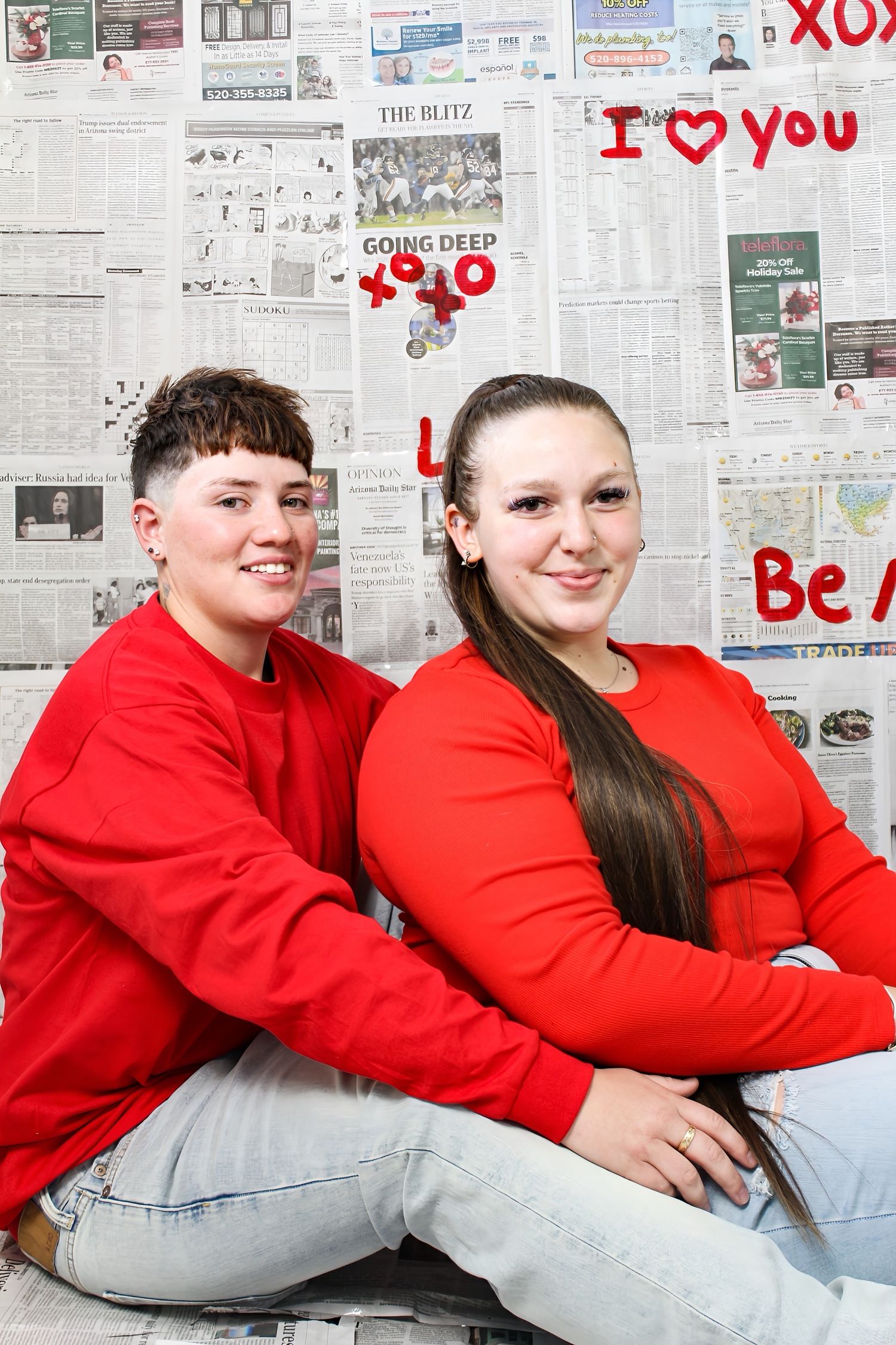 Two young women in red sweaters and jeans sit back-to-back smiling before a Valentine's Day newspaper wall.