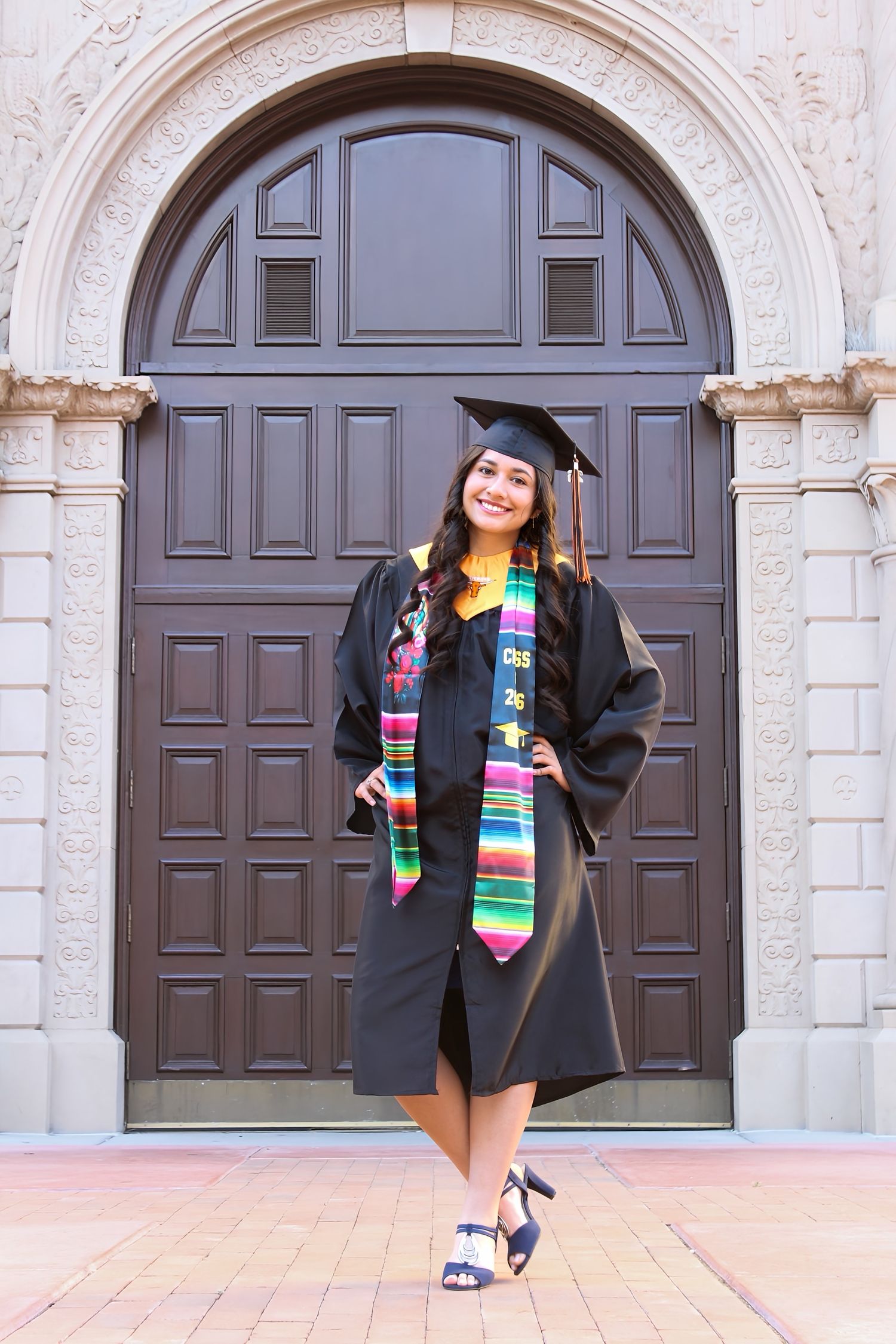 Happy female graduate in black cap and gown with colorful stole posing in front of an ornate arched doorway.