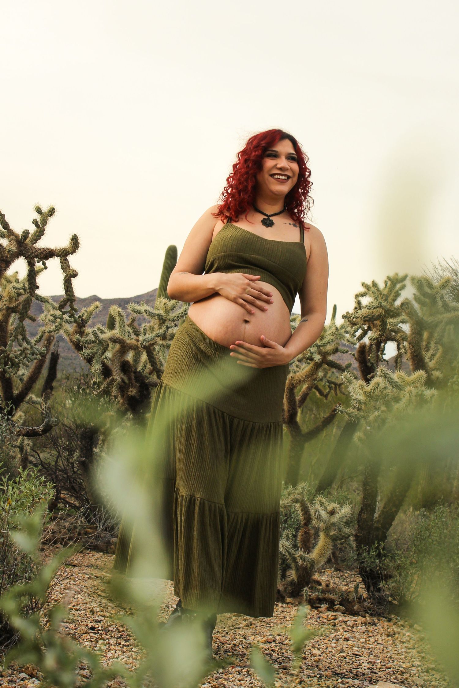 Joyful pregnant woman with red curly hair in olive dress cradling baby bump among desert cacti.