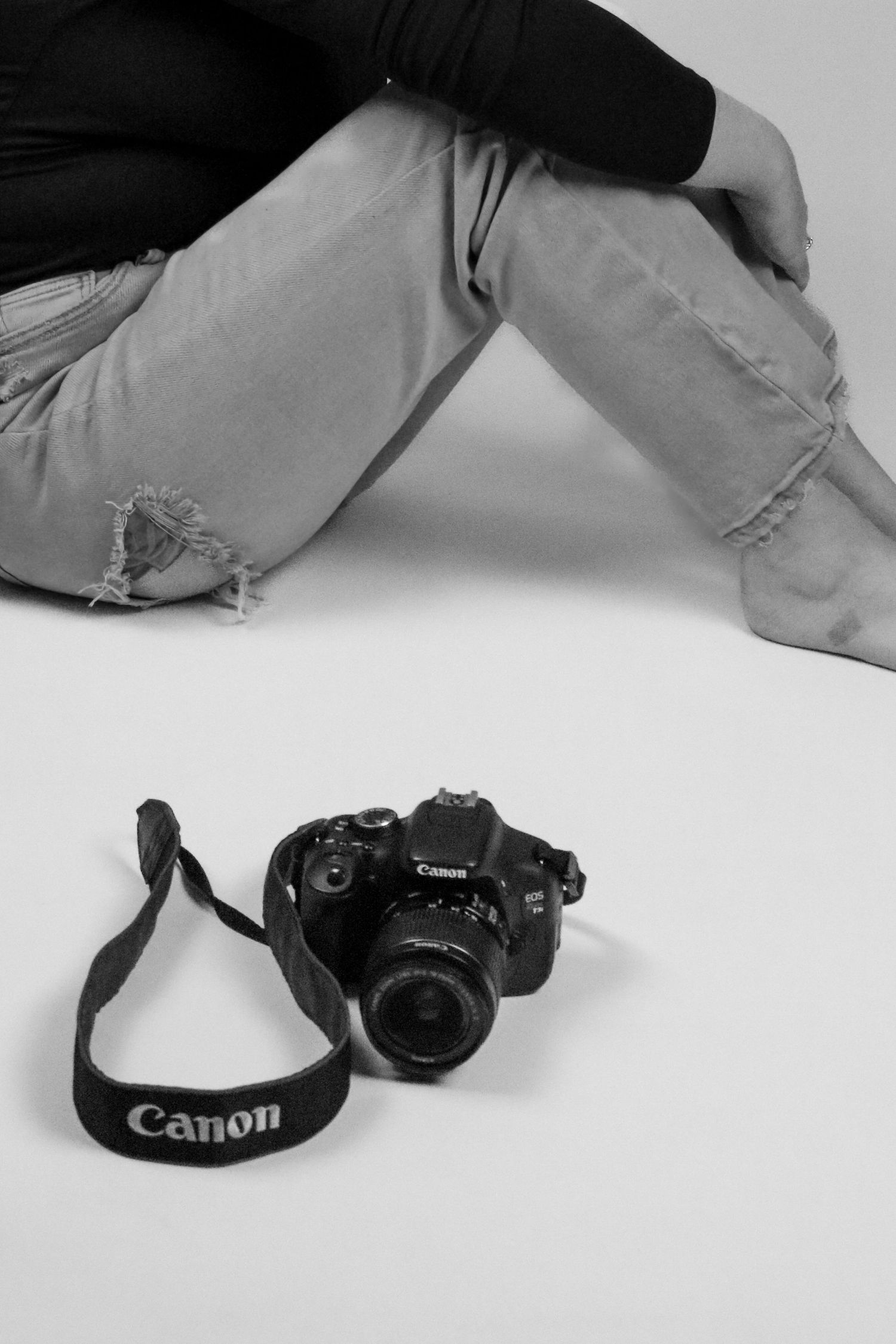 Black and white photo of Canon DSLR camera with strap on white floor next to seated person in jeans.