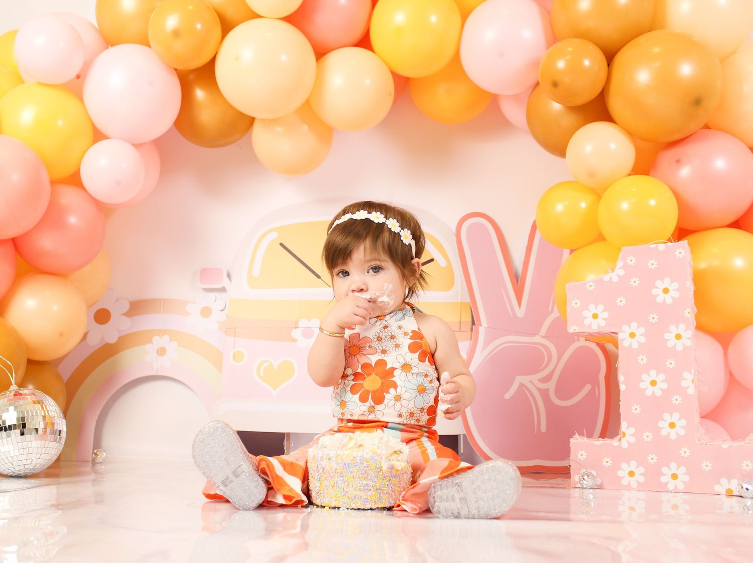 Baby girl celebrating first birthday with cake smash surrounded by pink and yellow balloon arch.