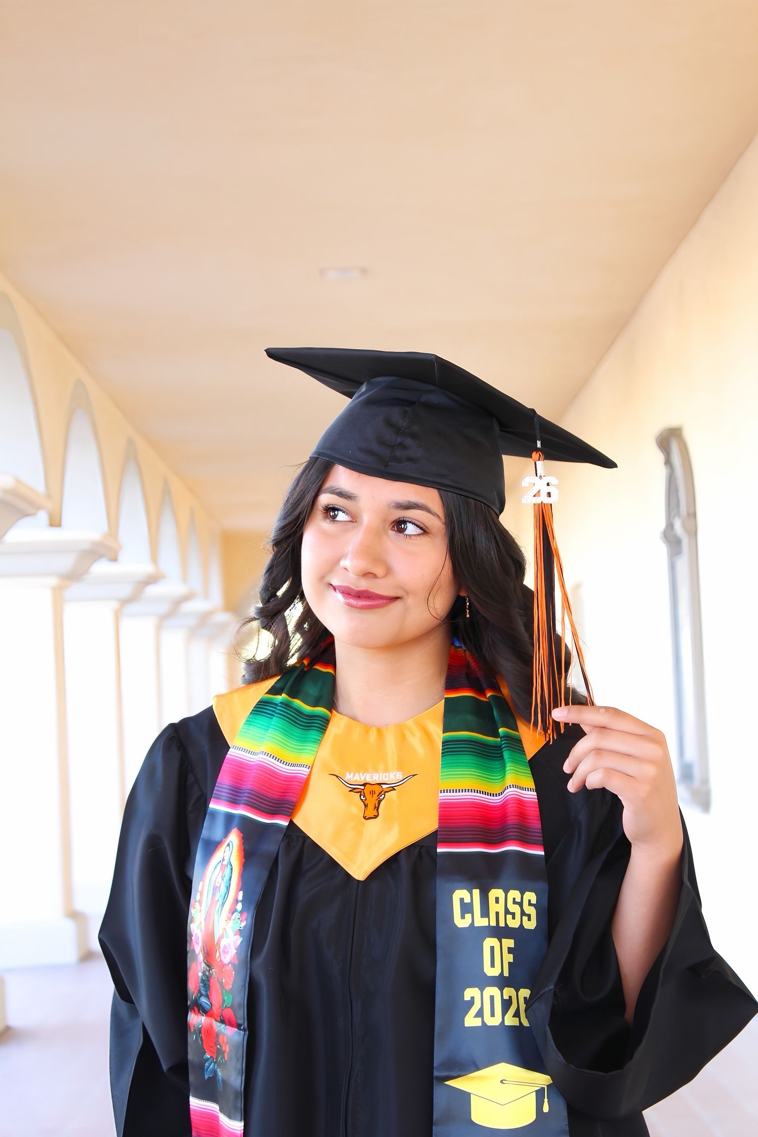 Female graduate in black cap and gown with colorful serape stole and Class of 2020 sash in arched corridor.