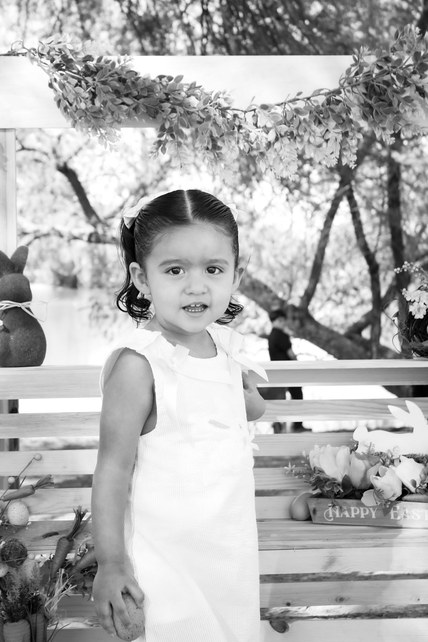 Black and white portrait of a young girl in a white dress standing at an Easter-themed outdoor backdrop.