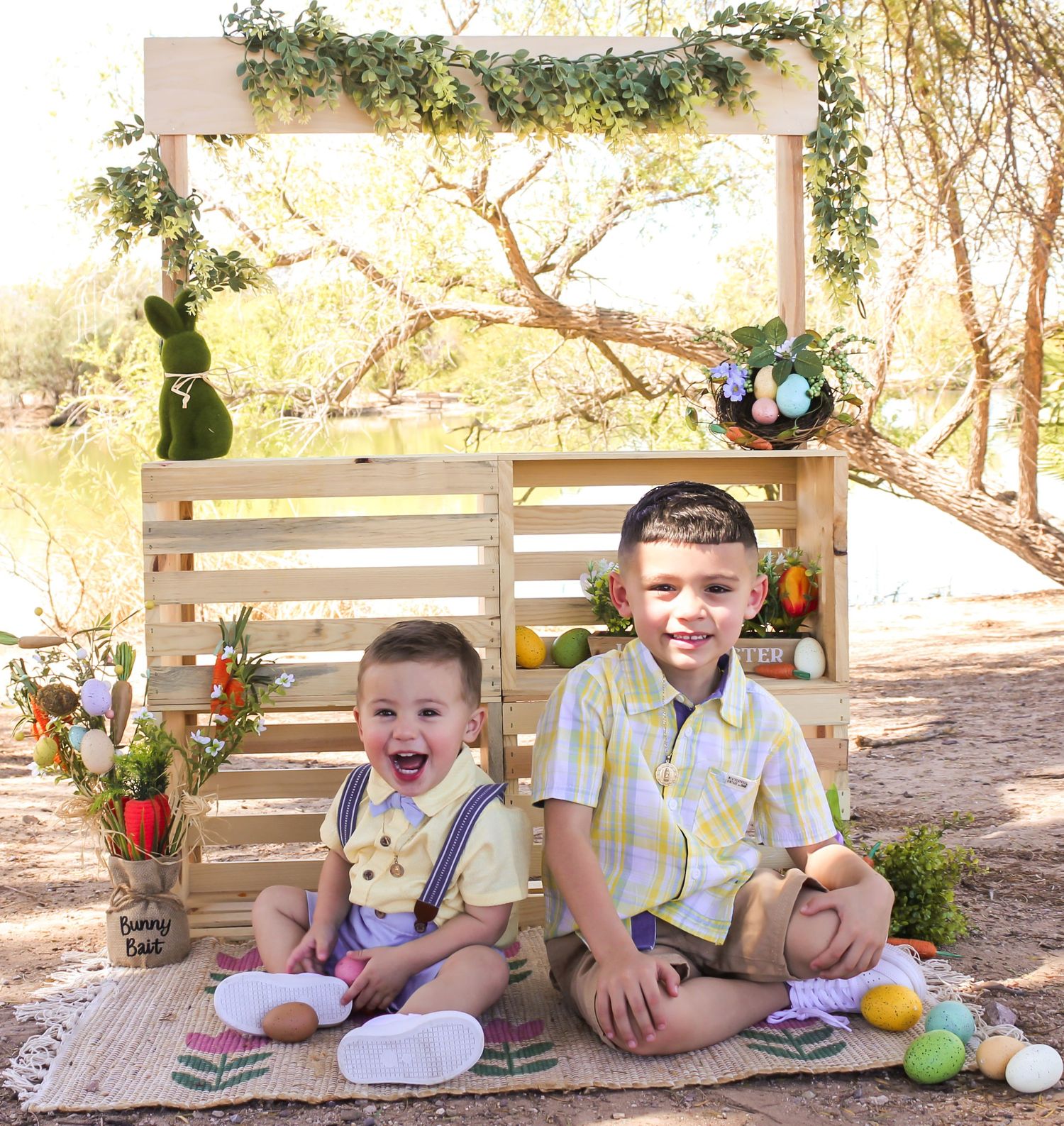 Two young brothers sitting together at an Easter-themed outdoor photo setup with colorful eggs and decorations.