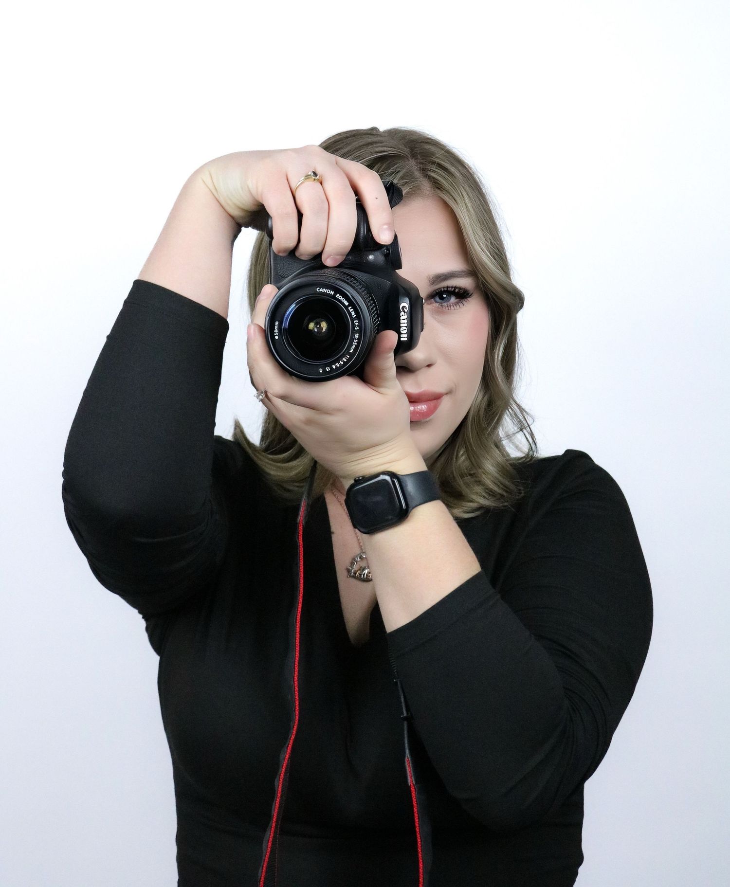 Female photographer in black top aiming Canon DSLR camera toward viewer with tattoo visible on wrist.