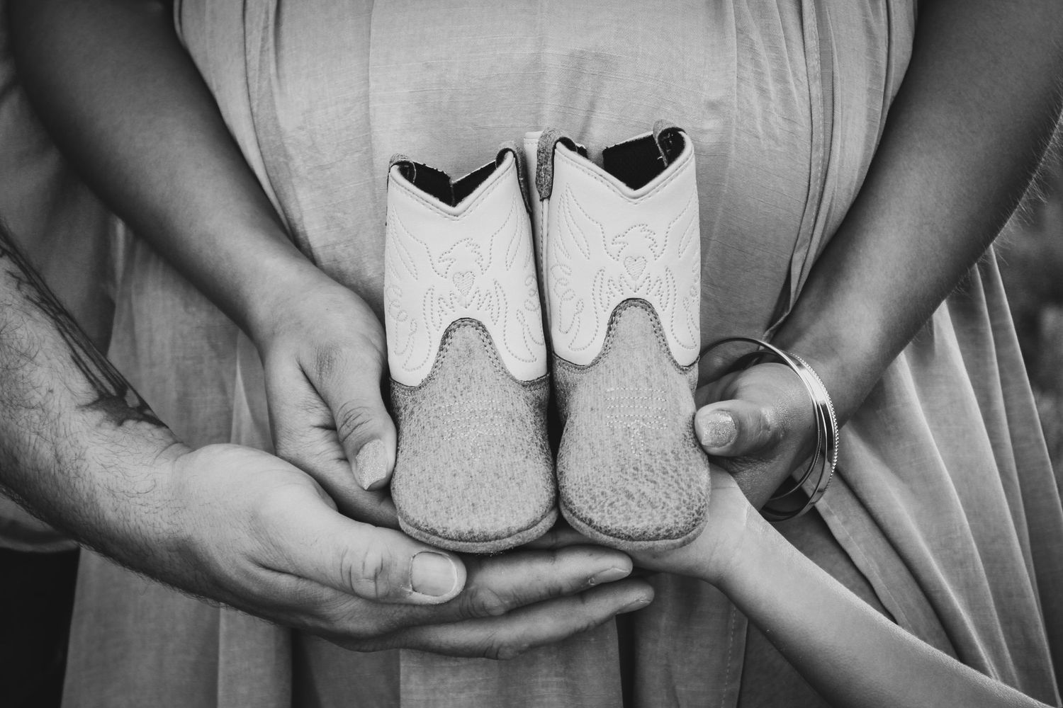 Black and white photo of couple holding tiny baby booties in front of pregnant belly outdoors.
