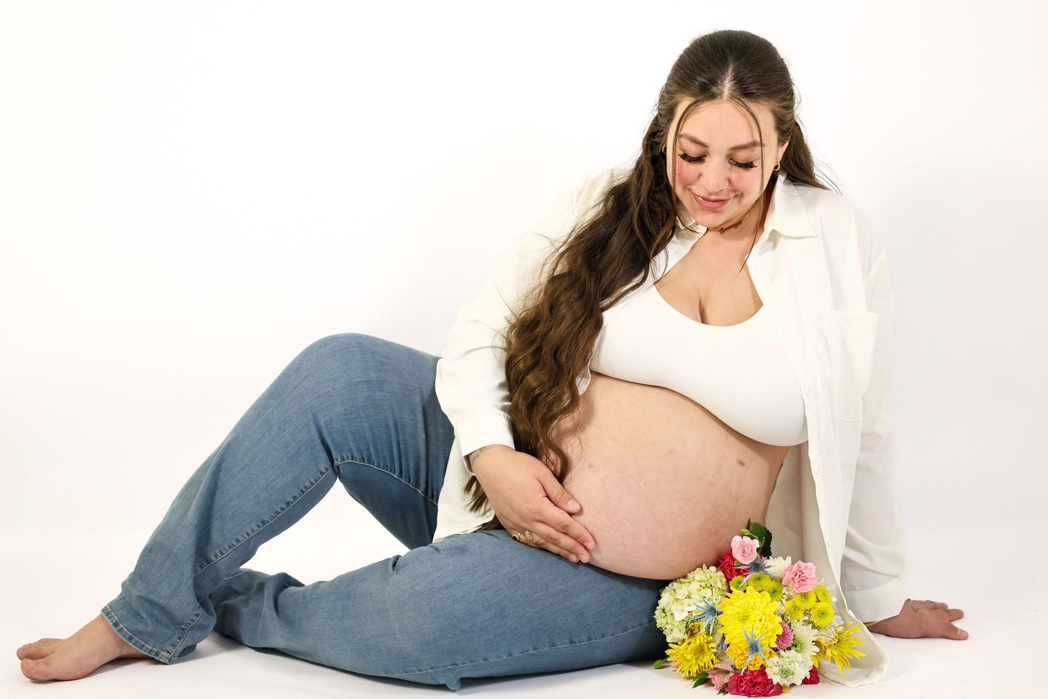 Pregnant woman in jeans and white blazer sits holding baby bump with flowers beside her.