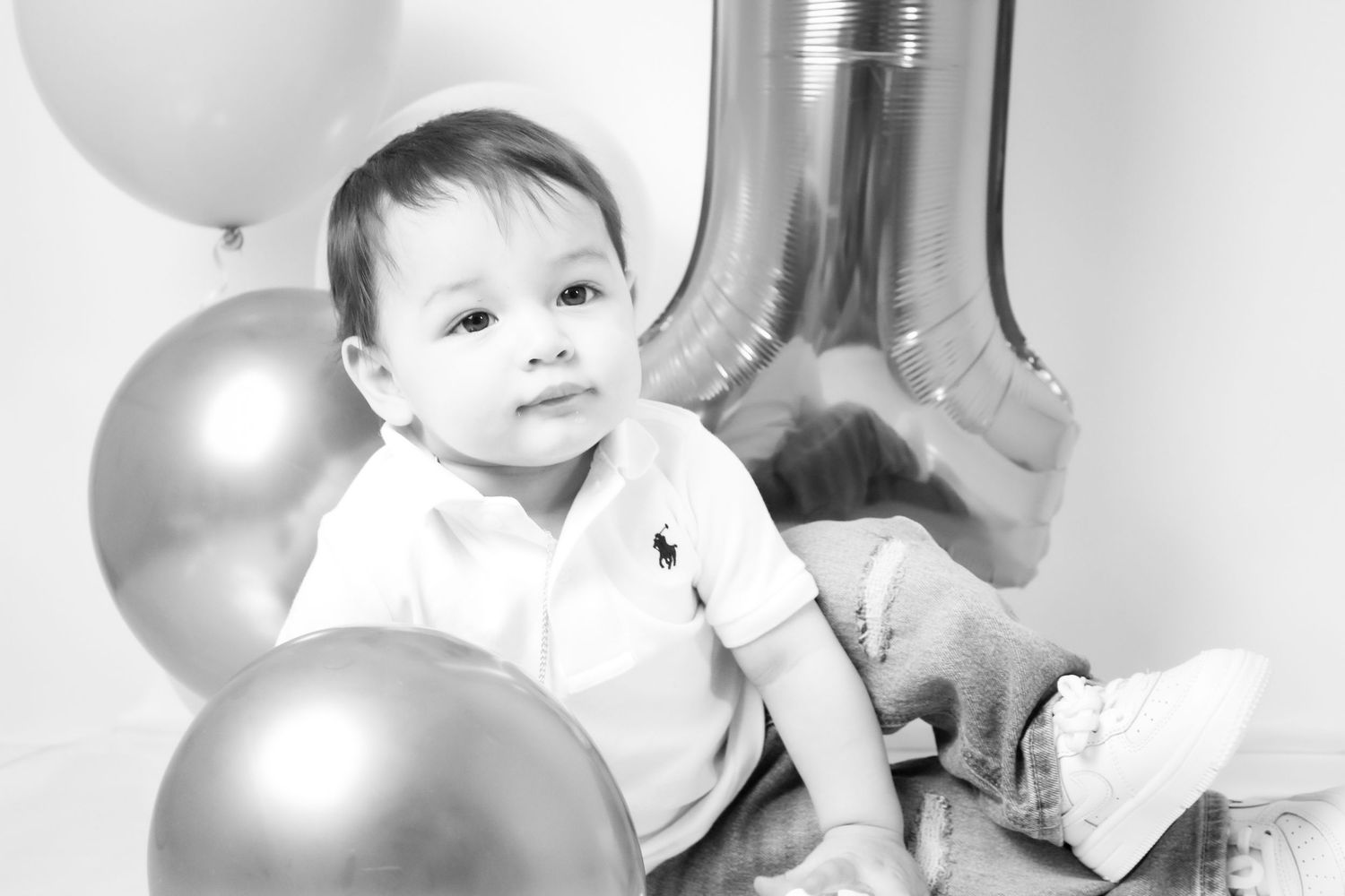 Black and white portrait of a baby boy sitting with metallic balloons at a first birthday celebration.