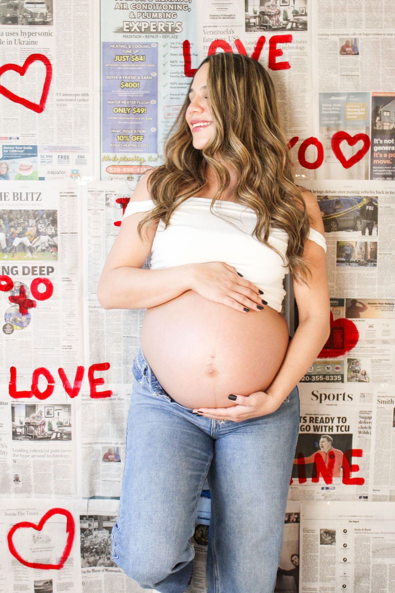 Smiling pregnant woman in white top and jeans with love-themed newspaper backdrop.