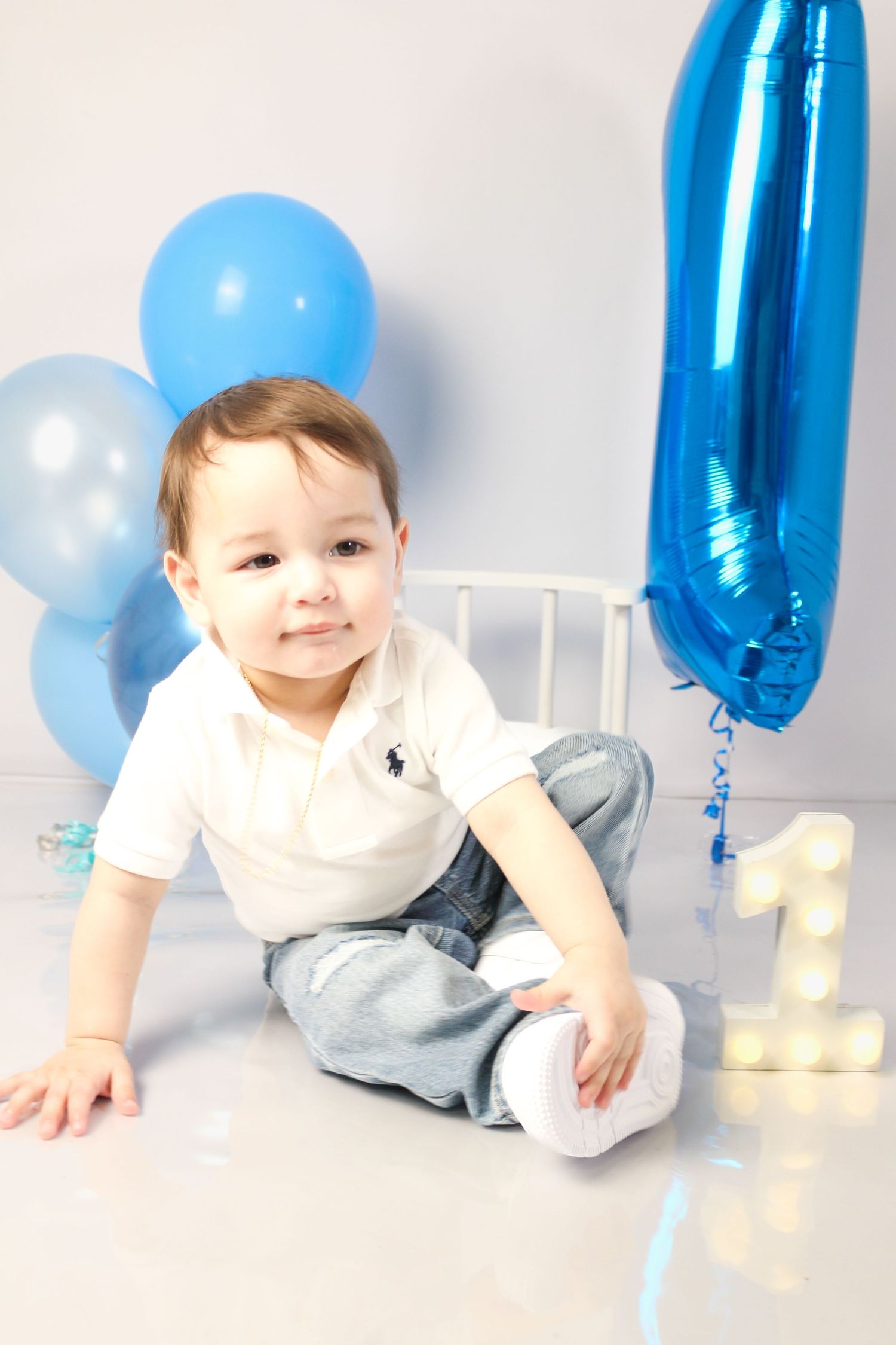 Smiling baby boy sitting on floor with blue balloons and number 1 decorations at his first birthday party.