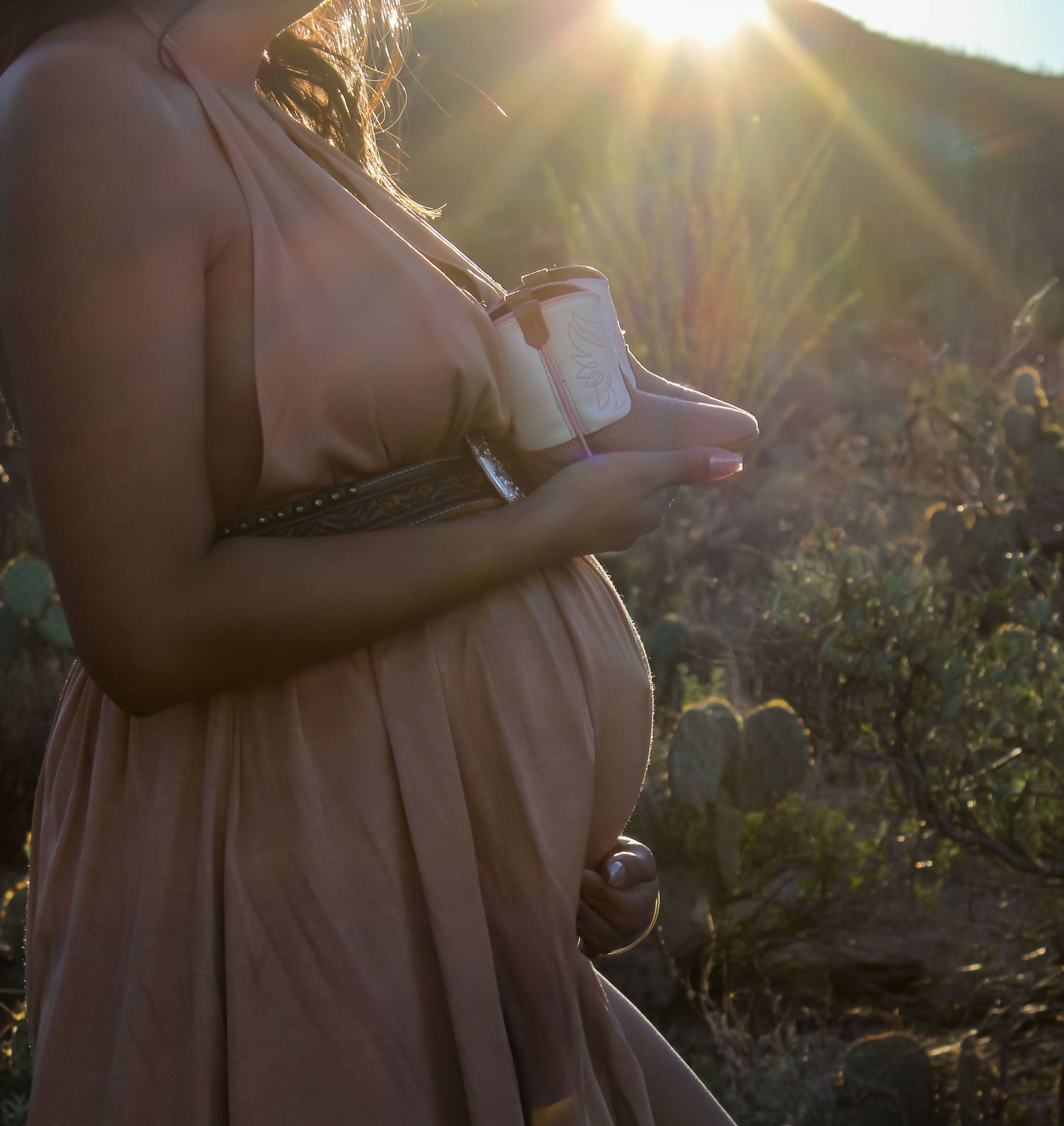 Pregnant woman in mauve dress holding baby shoes in golden sunlight in desert setting.