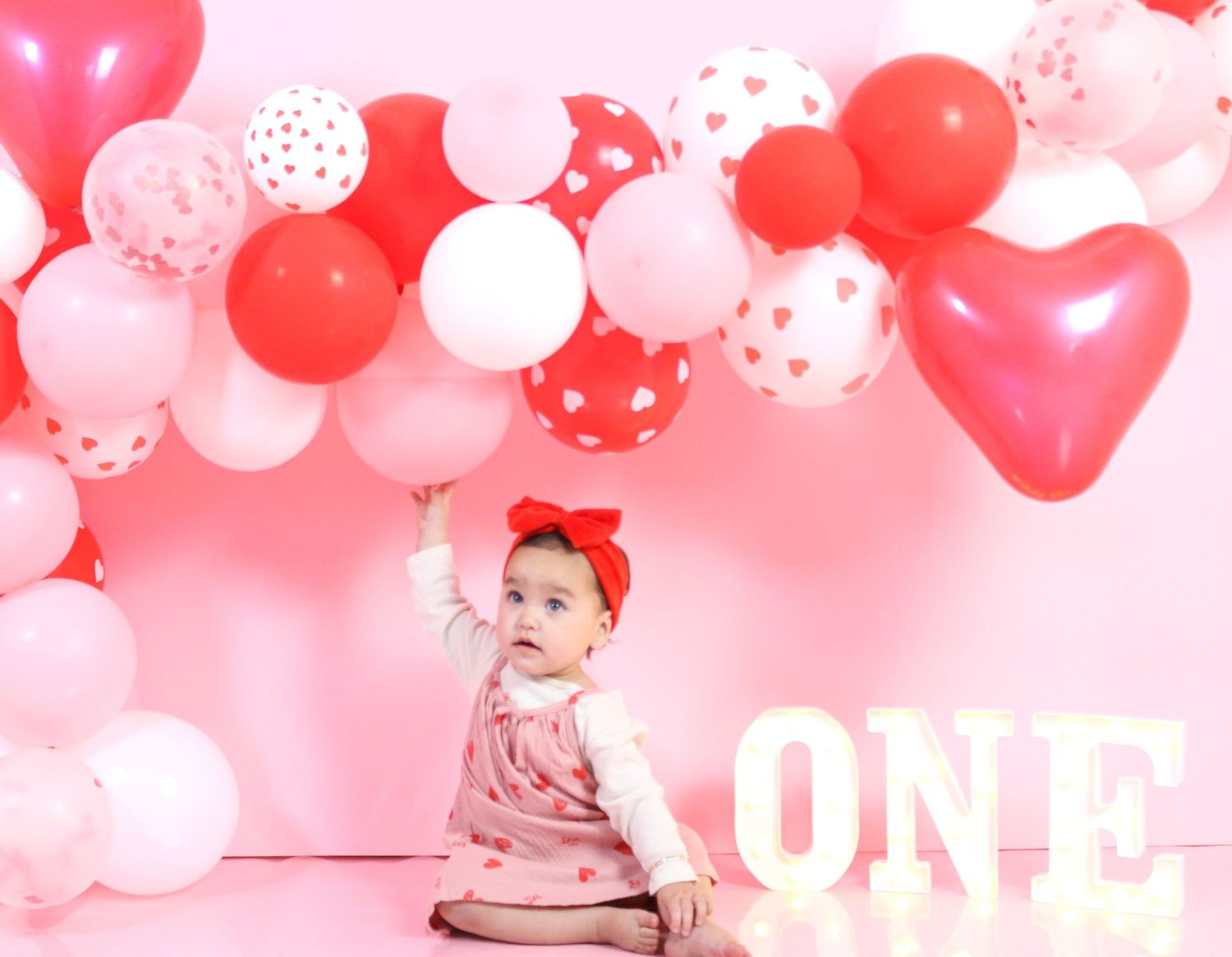 Baby girl in pink dress sits near ONE sign with red and pink heart balloon garland backdrop.