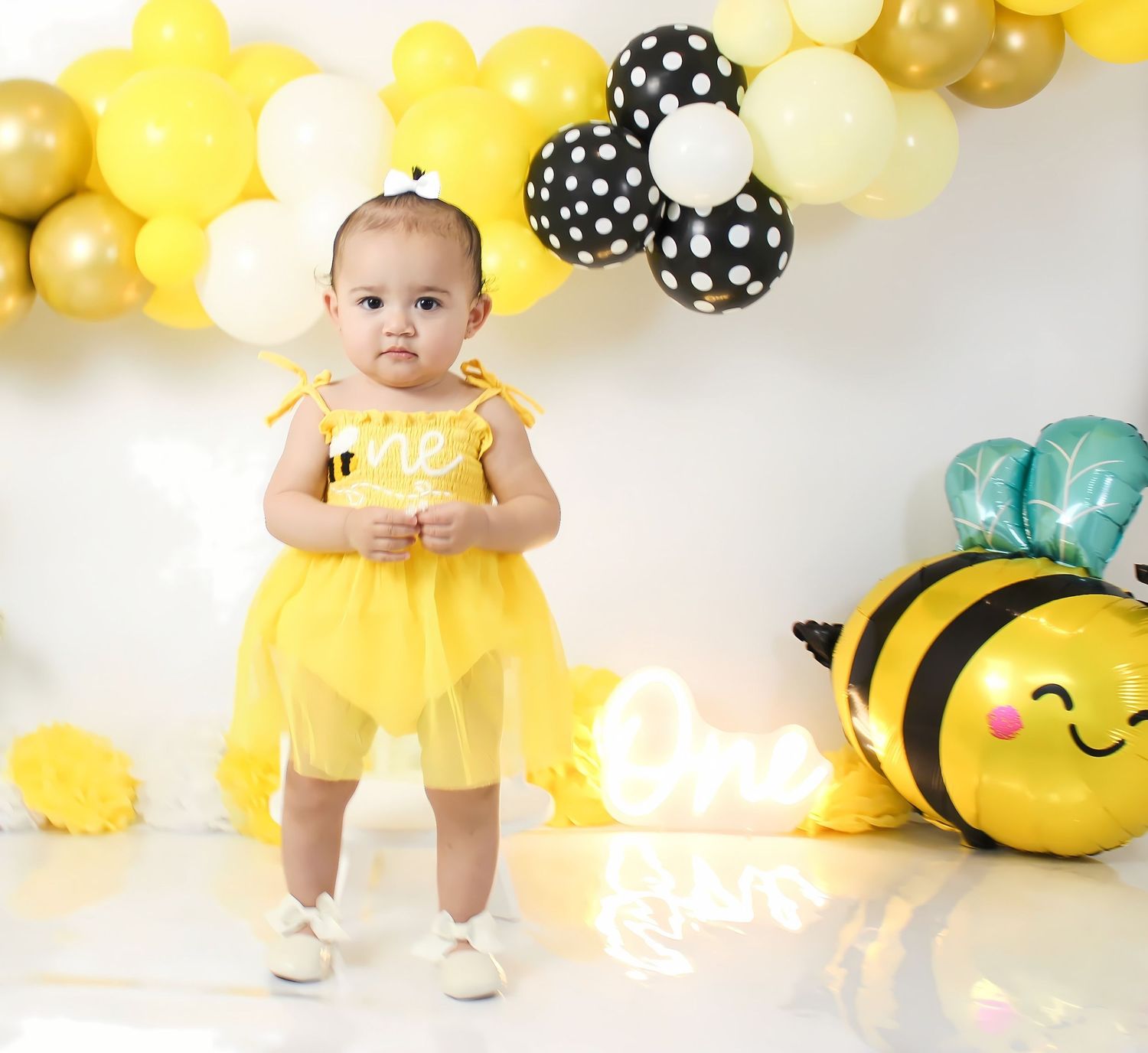 Baby girl in yellow bee costume celebrating first birthday with balloon garland and bee decorations.