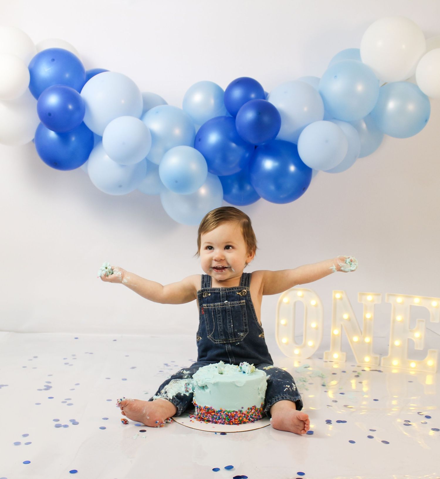 Happy baby boy in overalls celebrates first birthday with blue balloon garland and ONE marquee sign.