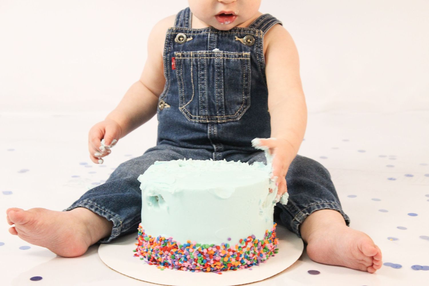 Baby boy in denim overalls smashing light blue sprinkle cake at first birthday cake smash session.