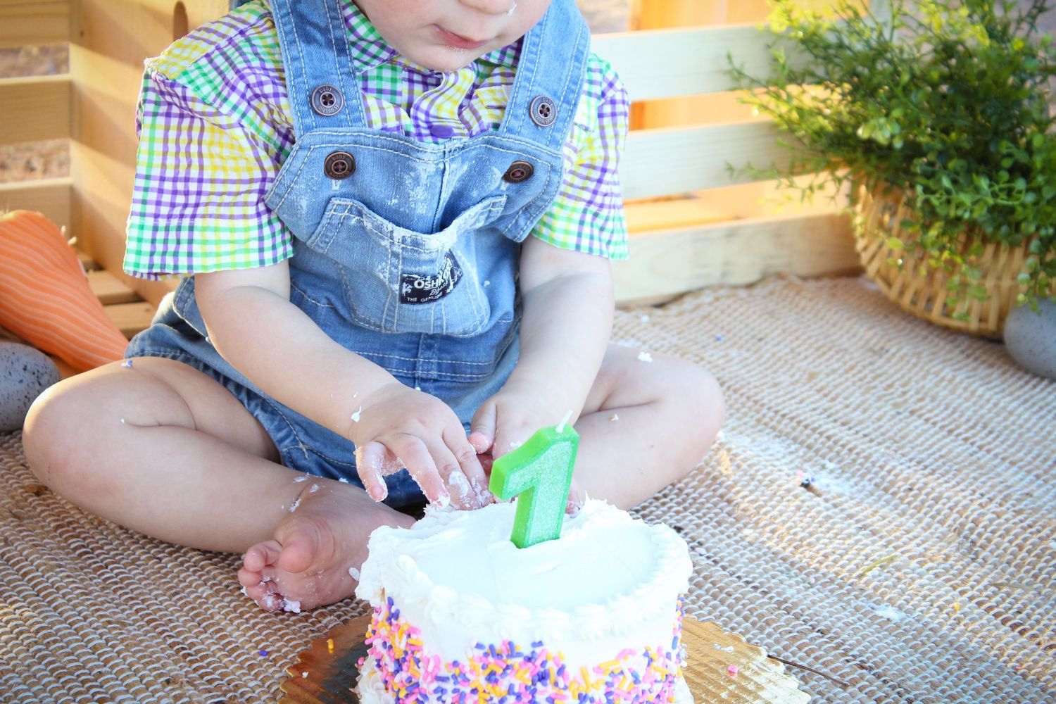 Toddler boy in denim overalls smashing a white sprinkle birthday cake with a number 1 green candle.