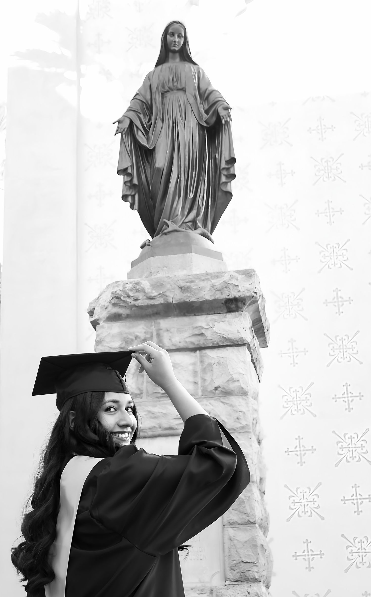 Smiling female graduate in cap and gown posing in front of a Virgin Mary statue in black and white photo.