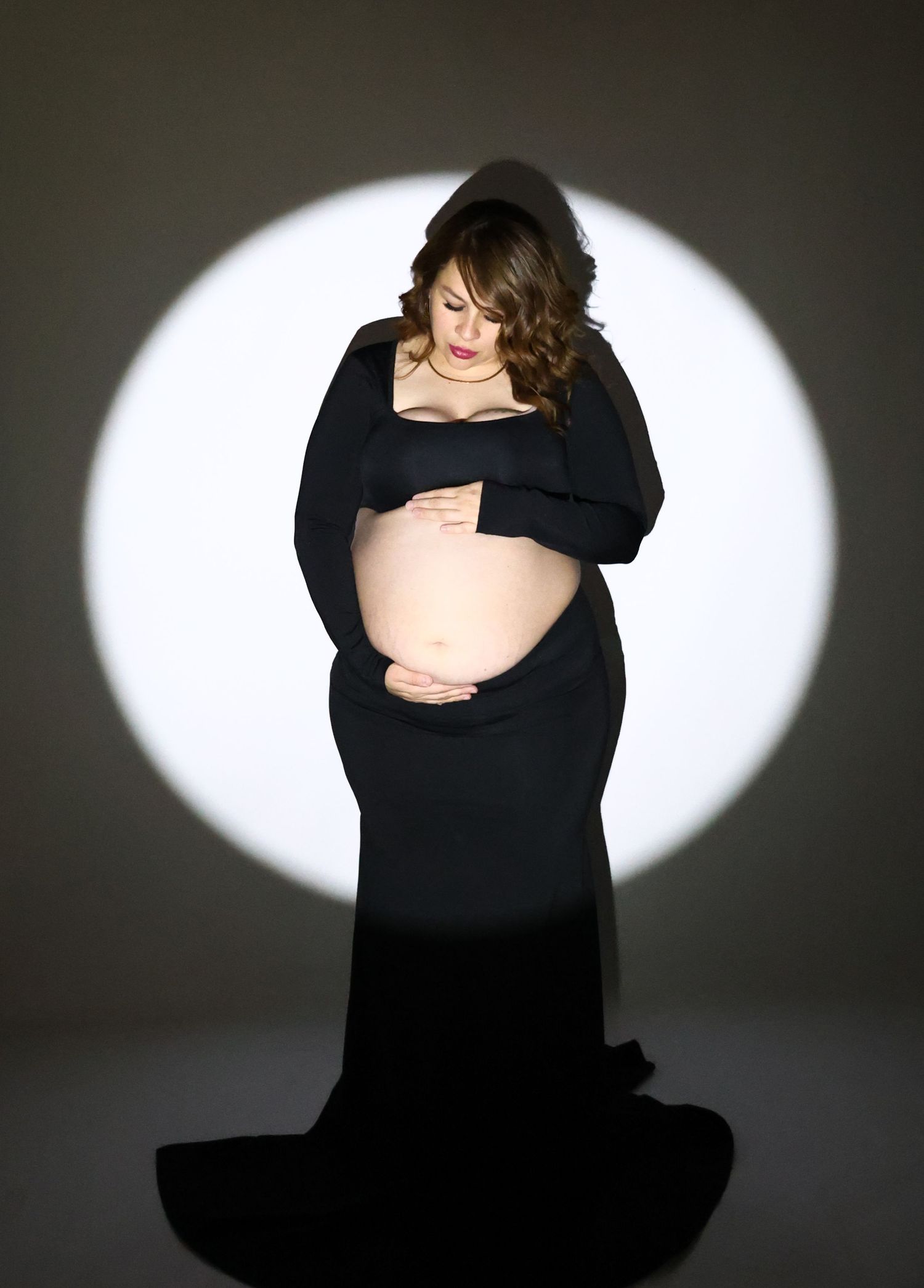 Pregnant woman in elegant black gown cradles baby bump against dramatic circular spotlight backdrop.