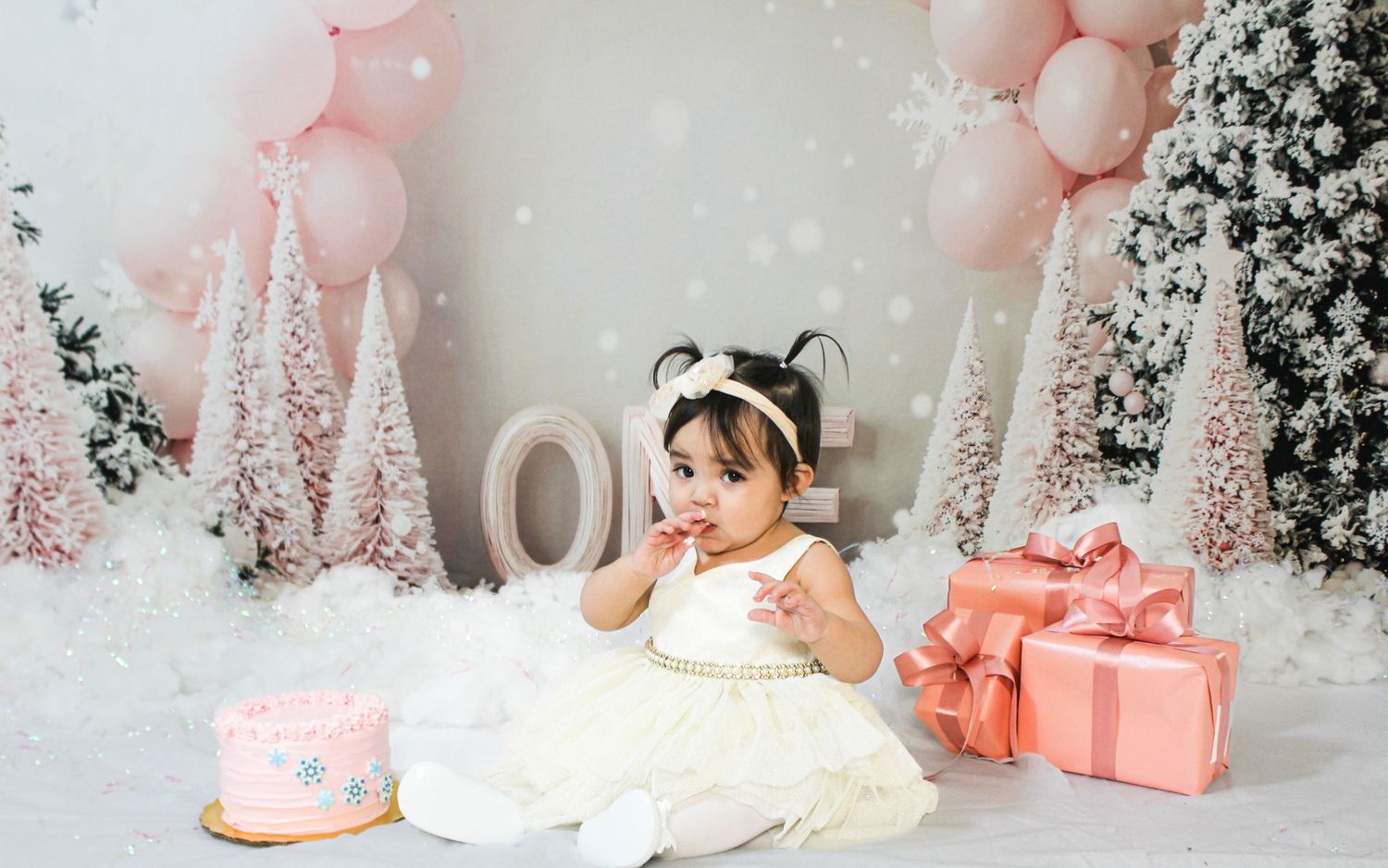 Baby girl in white dress at winter wonderland pink Christmas themed first birthday cake smash.
