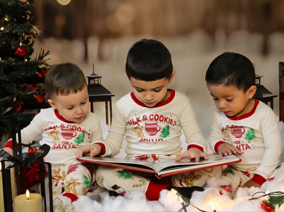Three young boys in matching Christmas pajamas sit together reading a book by a decorated tree.