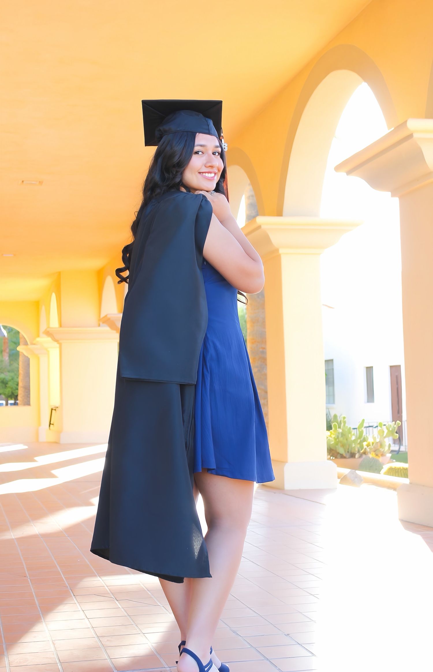 Young woman in blue dress and black graduation cap and gown smiling over her shoulder in an arched hallway.