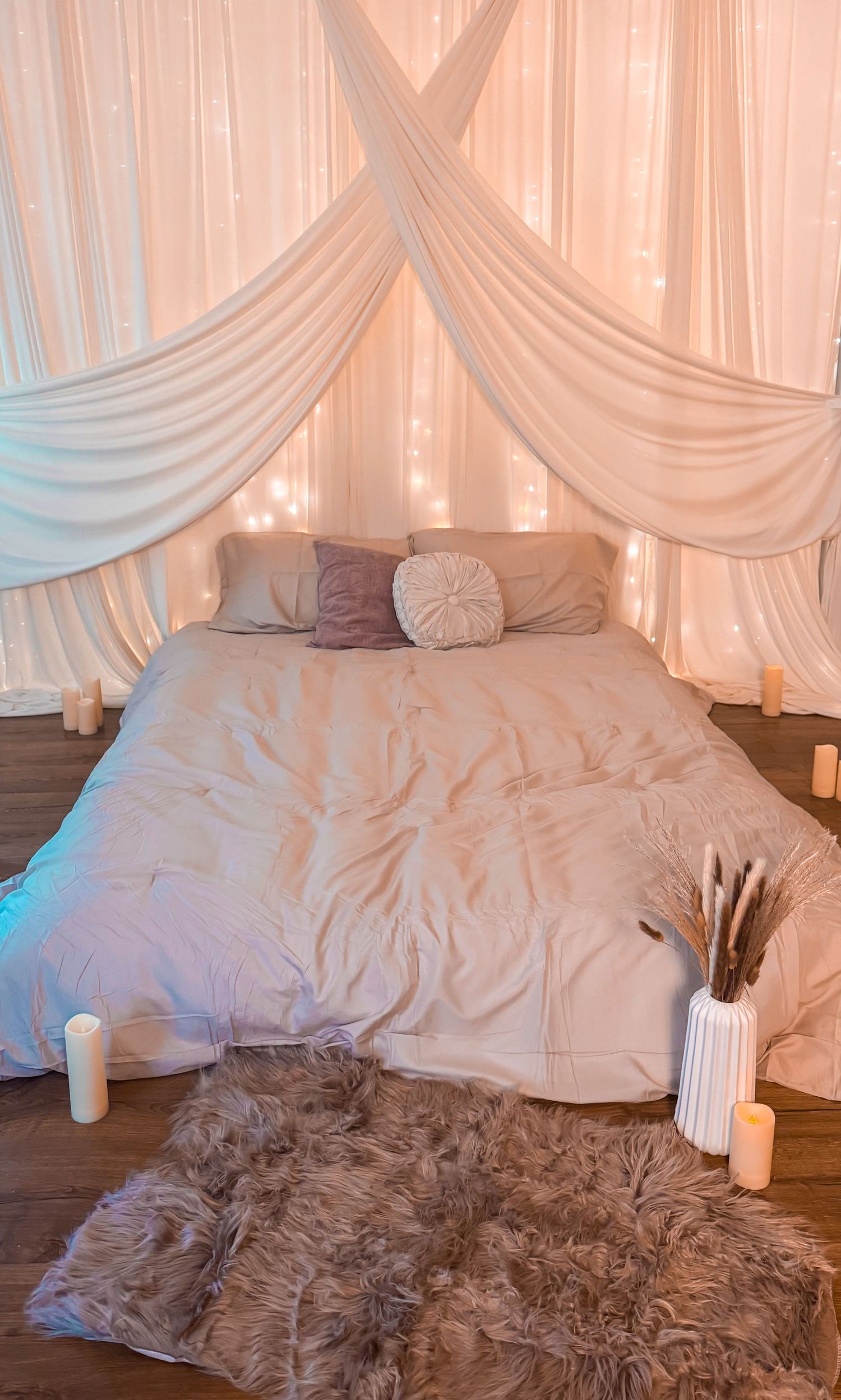 Romantic bedroom setup with white draped canopy, fairy lights, candles, fluffy rug, and neutral bedding on hardwood floor.
