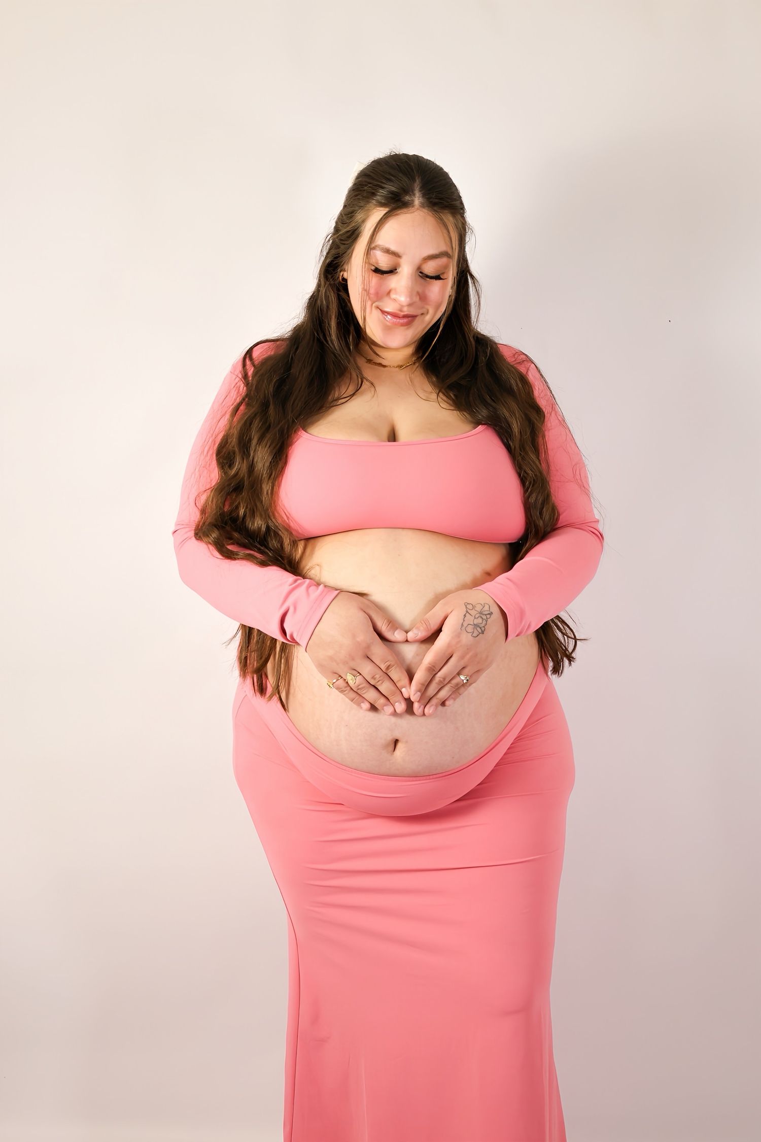 Pregnant woman in pink two-piece outfit makes heart shape with hands on baby bump.