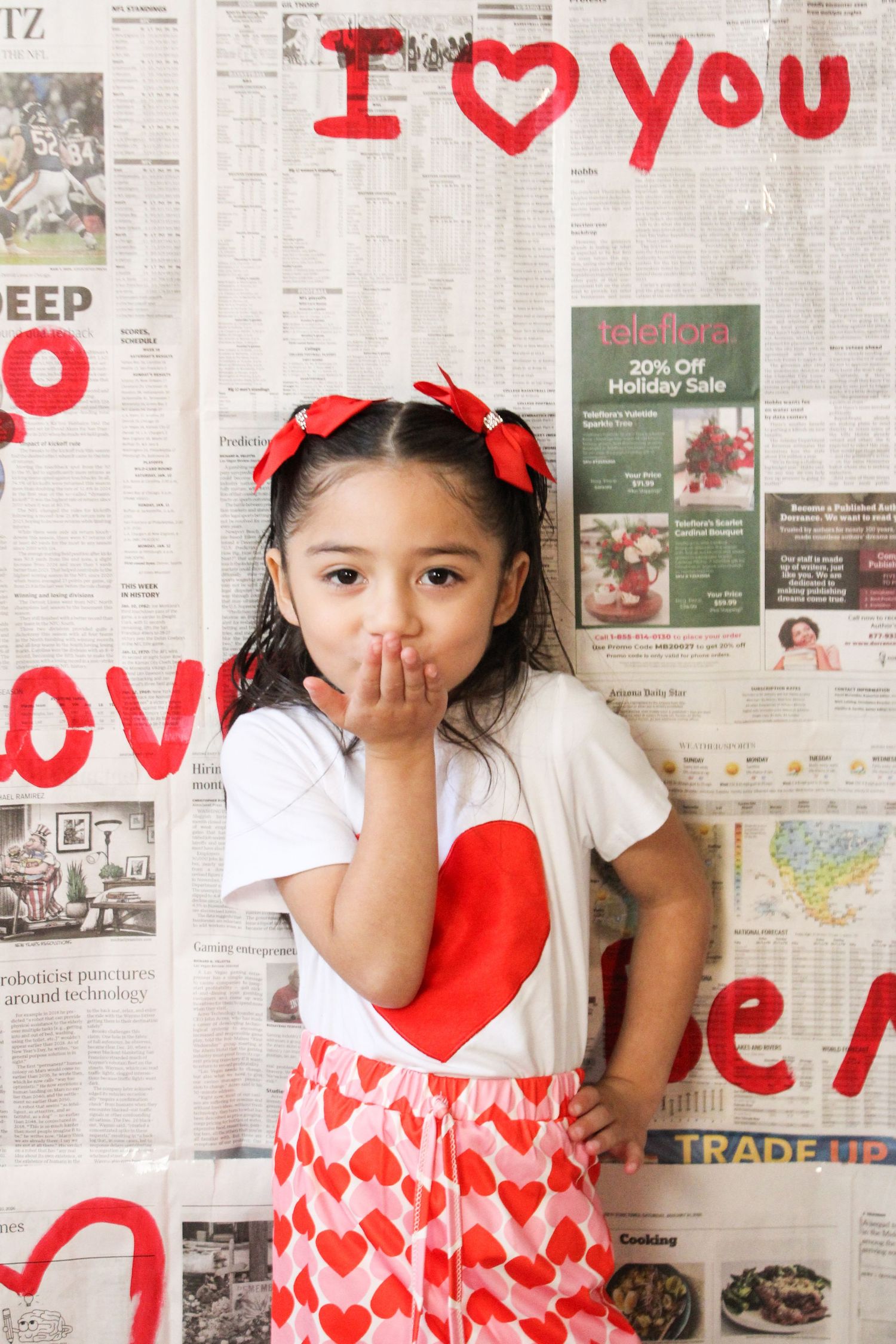 A young girl in a heart-print outfit blows a kiss in front of a Valentine's Day newspaper wall.