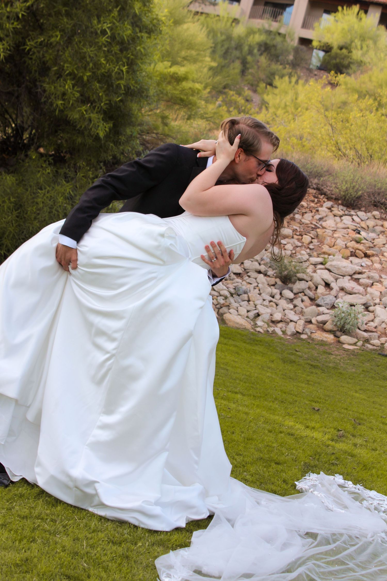Bride and groom sharing a romantic dip kiss outdoors in a lush garden setting on their wedding day.