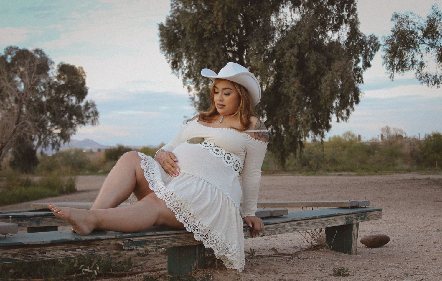 Pregnant woman in white dress and cowboy hat sitting outdoors at sunset.