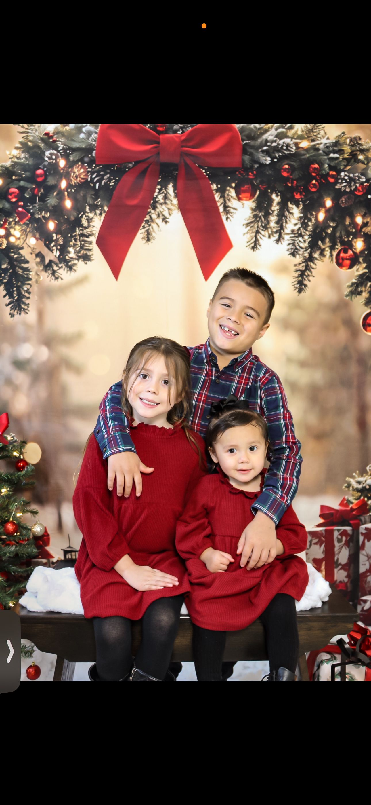 Three children in red holiday outfits pose together under a festive Christmas wreath and bow backdrop.