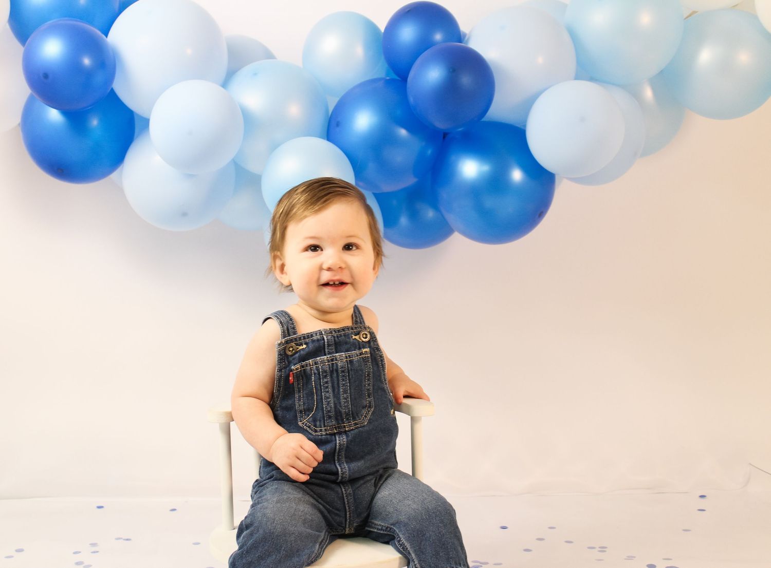Smiling baby boy in denim overalls sitting under blue balloon arch at first birthday session.