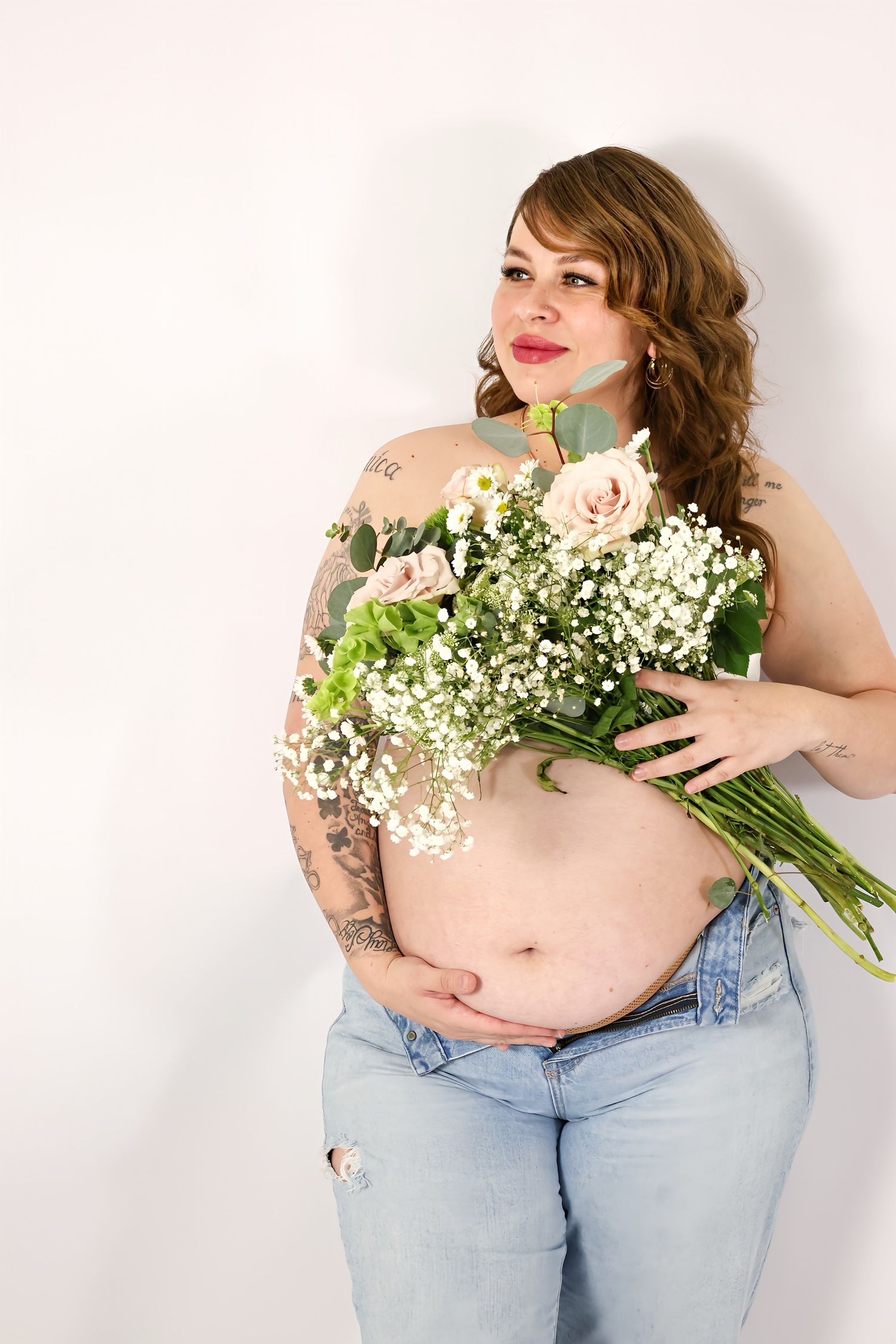 Tattooed pregnant woman in ripped jeans holds bouquet of roses and baby's breath flowers.