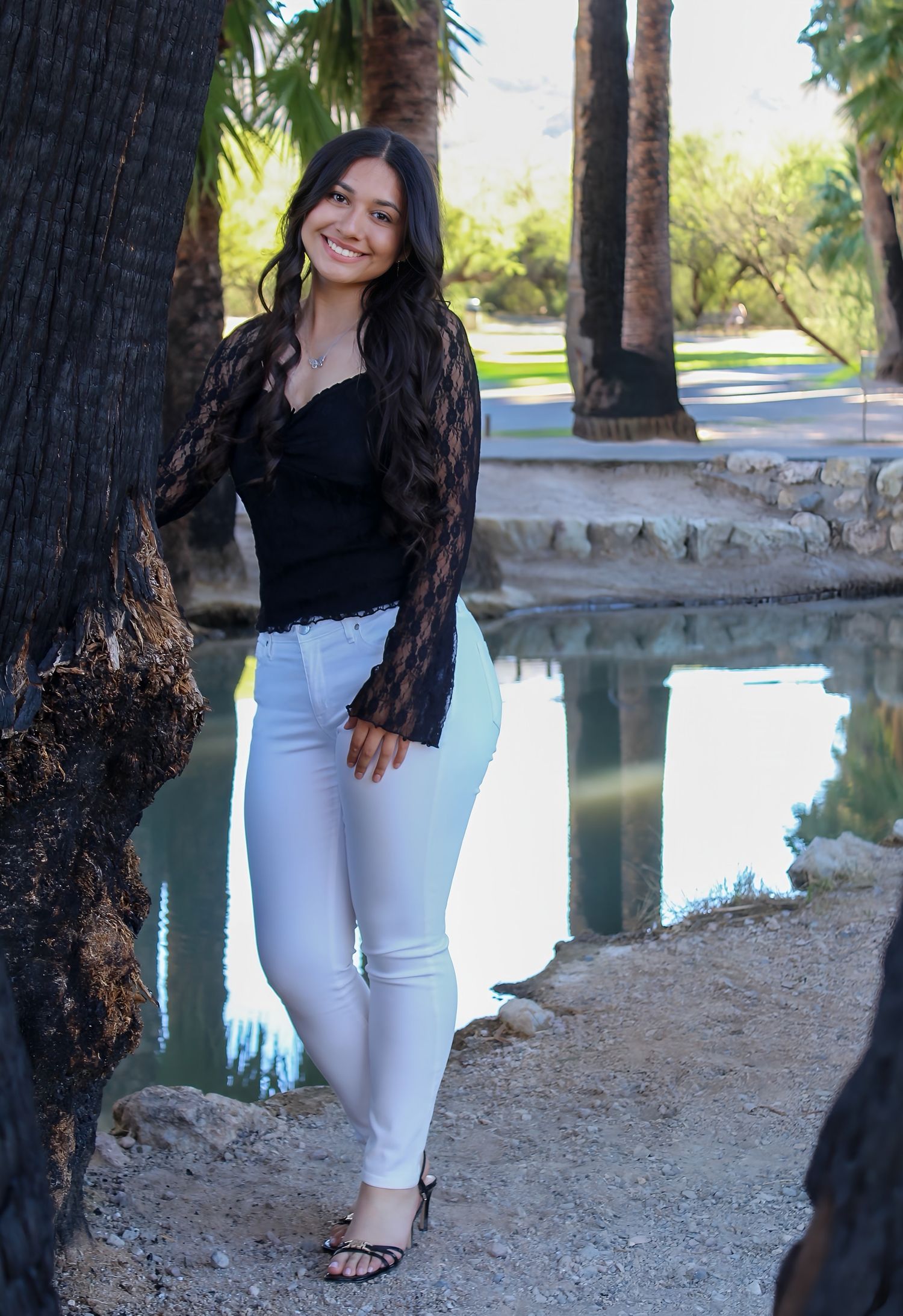 Young woman in black lace top and white jeans smiling beside a tree near a reflective pond in a park.