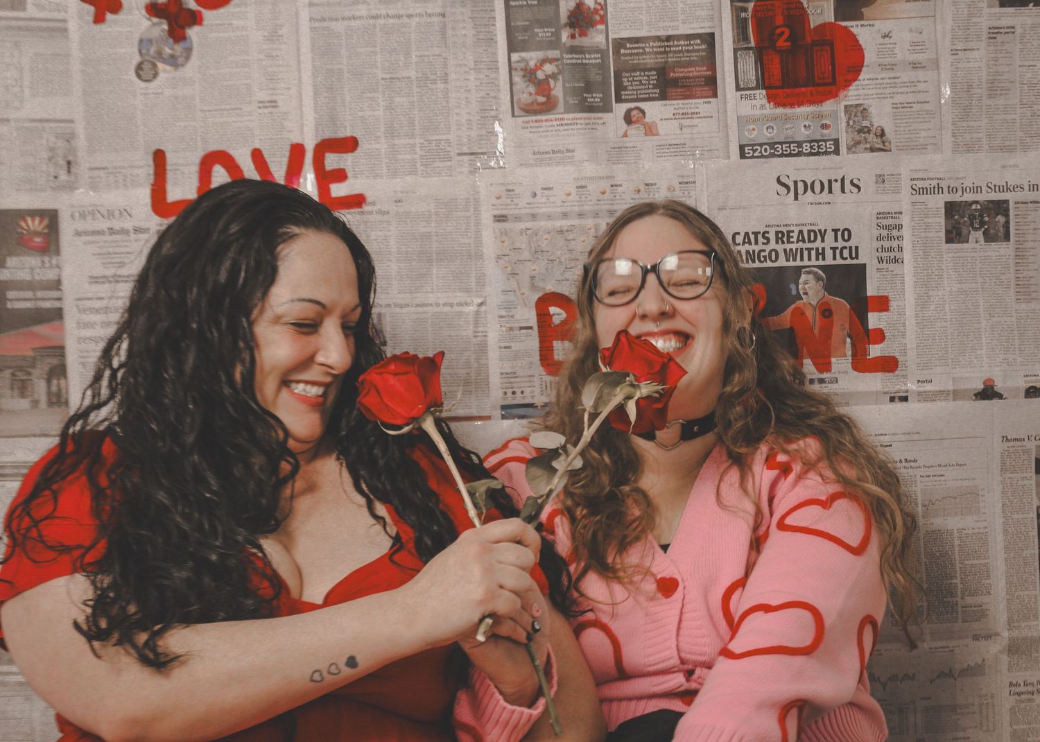 Two women laugh and smell red roses together in front of a Valentine's Day newspaper backdrop.