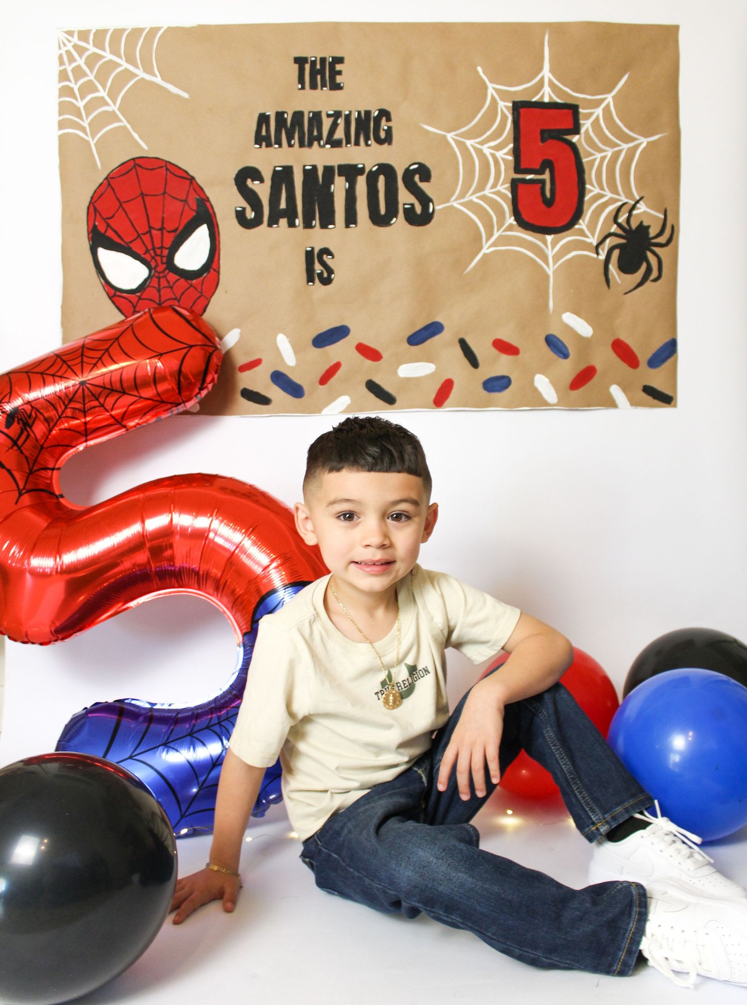 Boy named Santos celebrates his 5th birthday with a Spiderman themed banner and colorful balloons.
