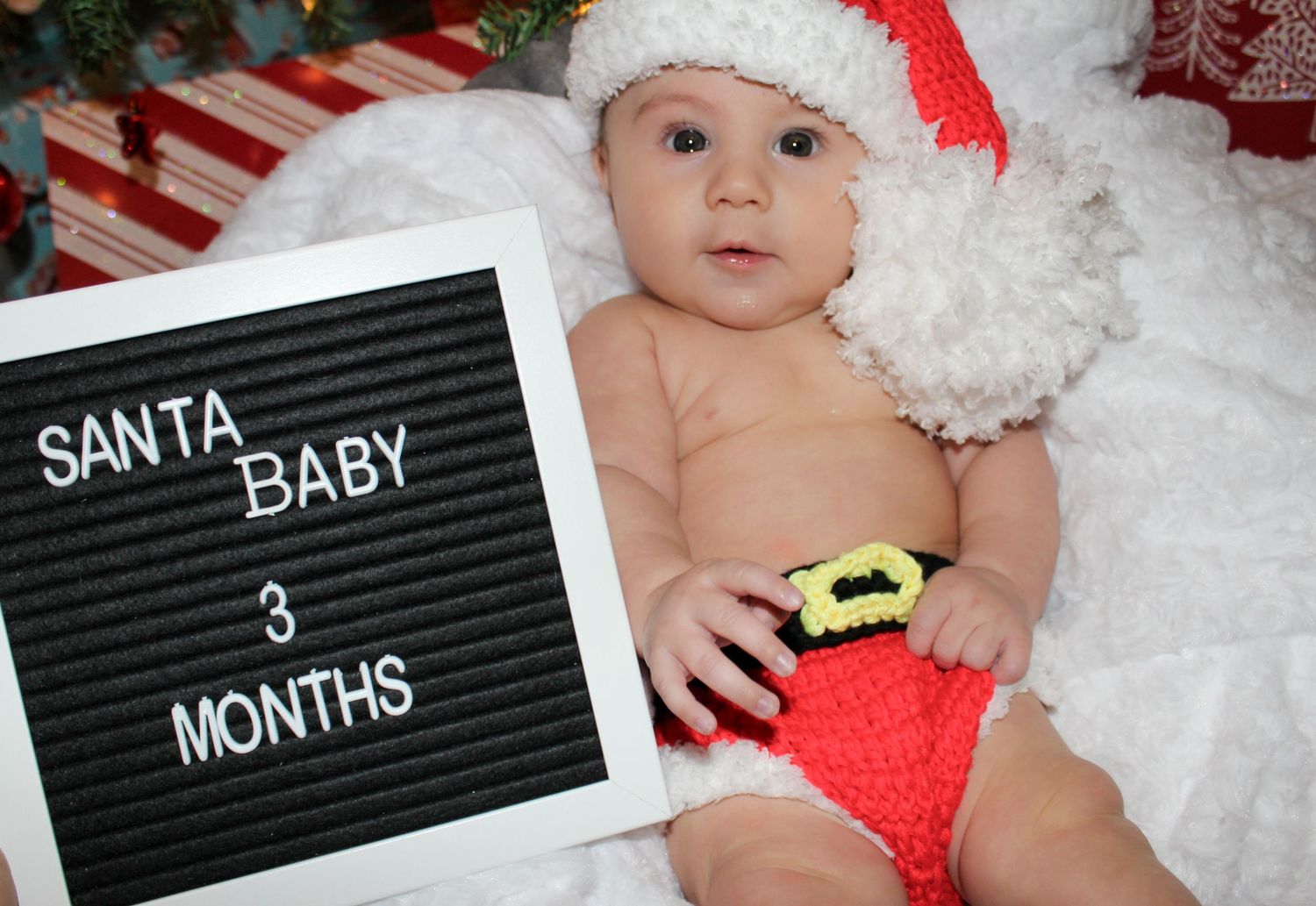 Three-month-old baby in Santa hat and outfit posed next to letter board for Christmas photo.